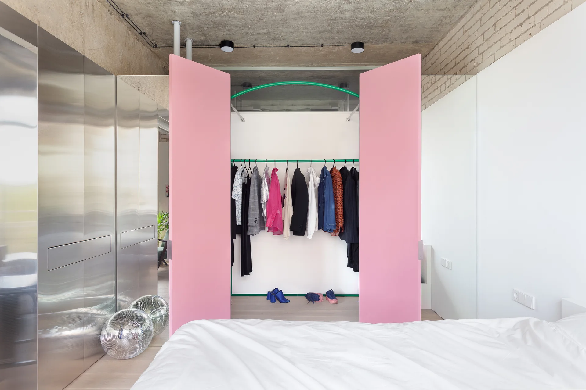 A bedroom features open pink closet doors, a green clothing rack, reflective metal cabinetry, and an exposed concrete ceiling.
