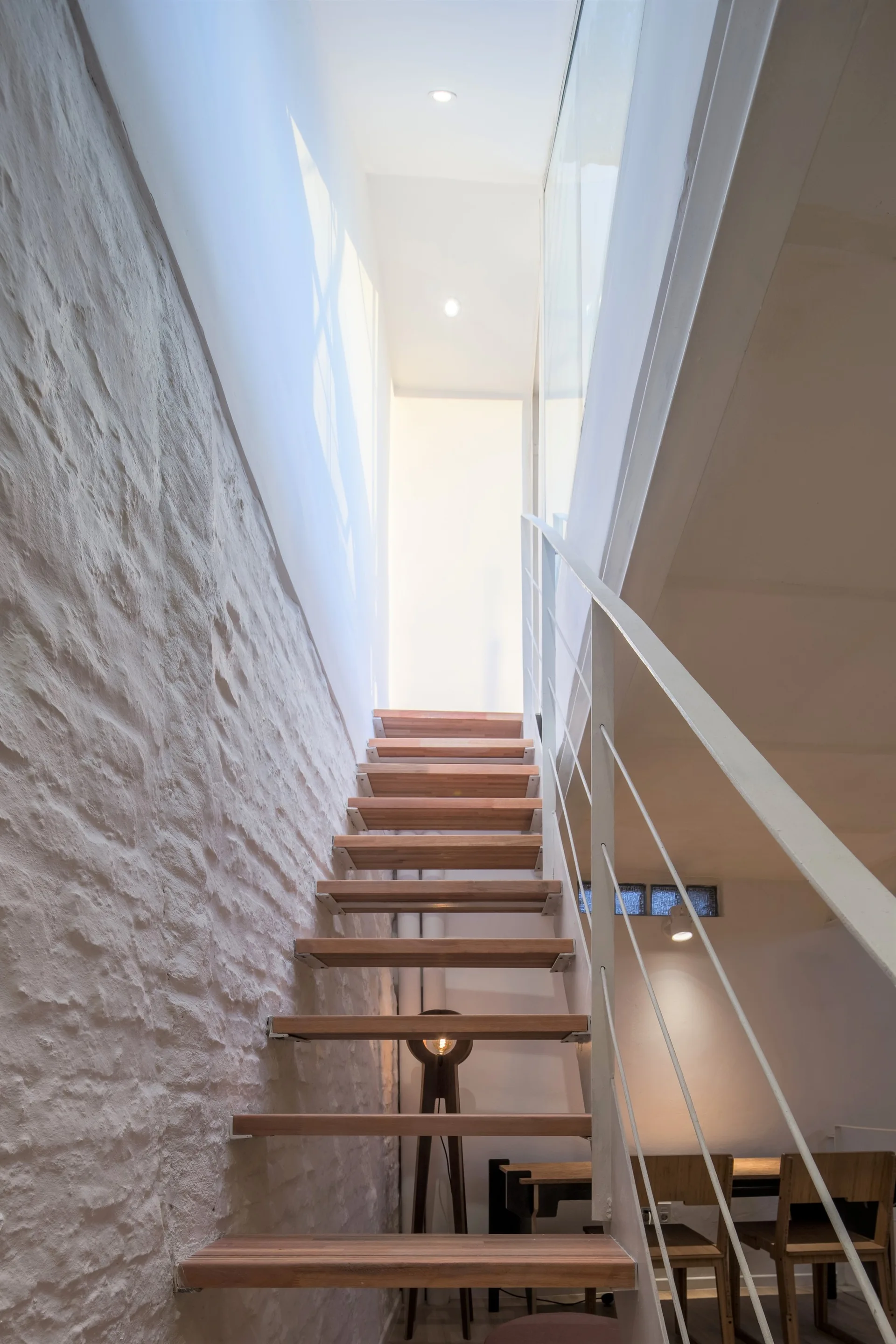 A modern floating staircase with natural wood treads and white metal railings ascends next to a rough white-painted stone wall.