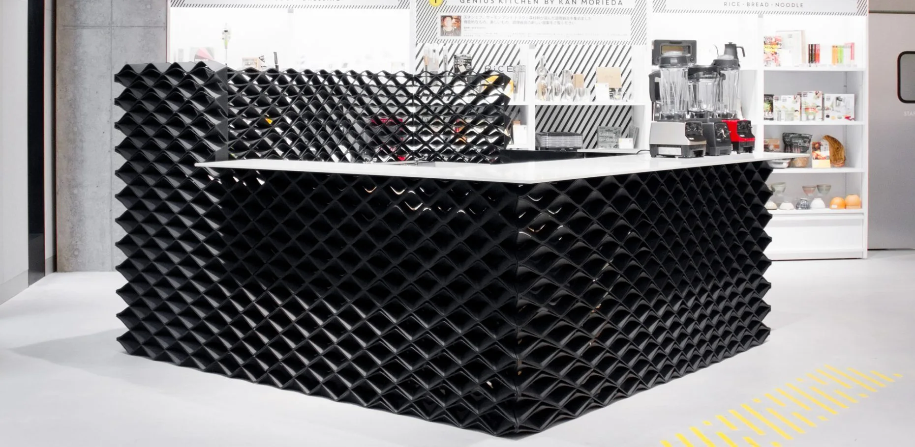 A modern kitchen island features a black 3D geometric relief facade and a smooth white countertop displaying several blenders.