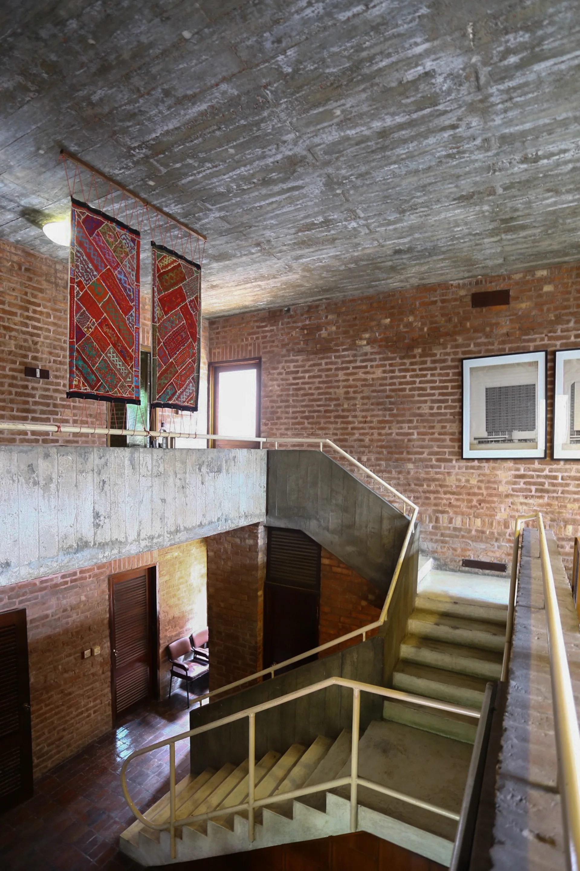 A multi-level concrete staircase with white metal railings is framed by exposed red brick walls, a rough concrete ceiling, and hung red tapestries.