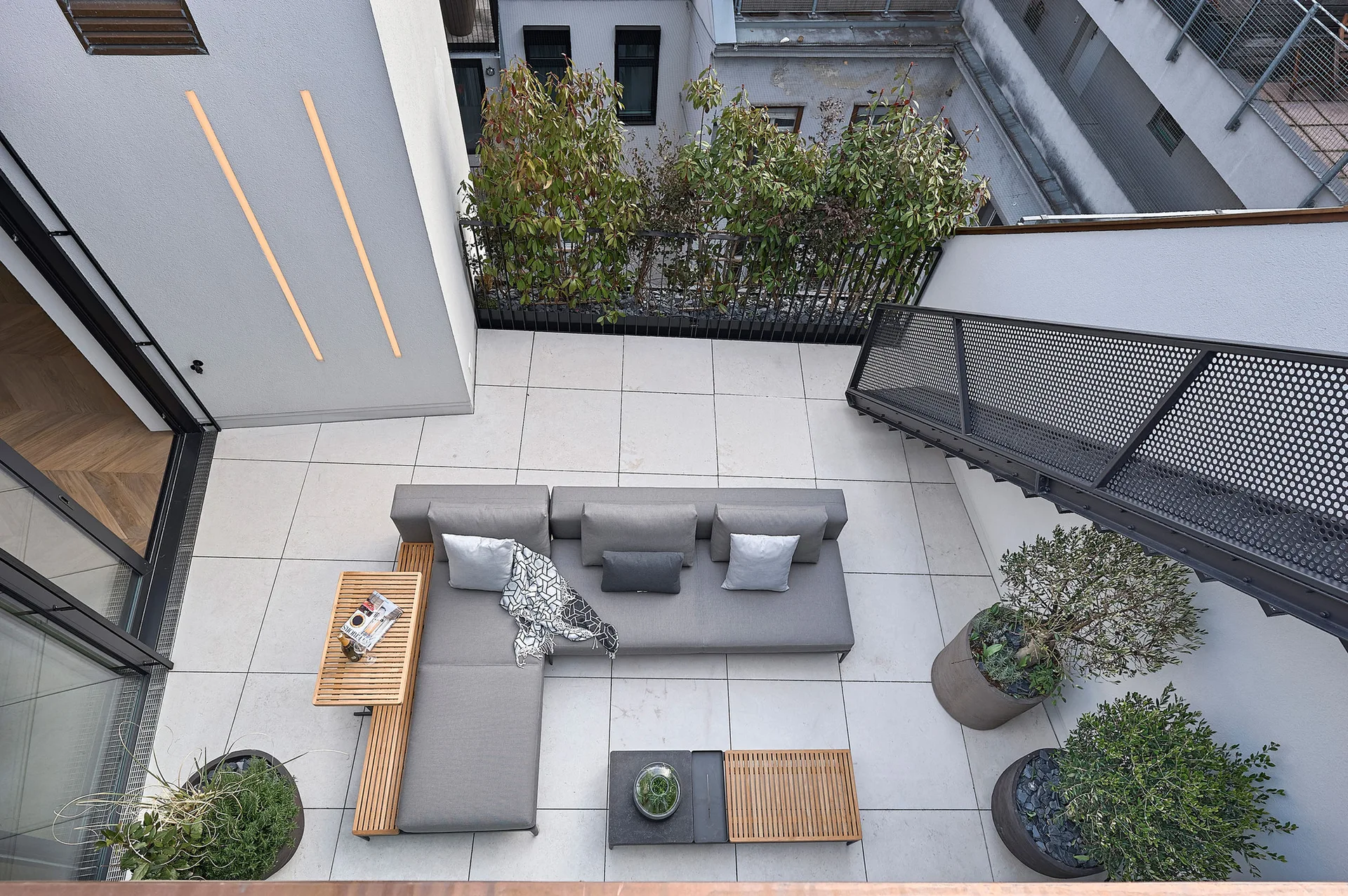 A high-angle view of a modern outdoor terrace featuring a grey sectional sofa, wood-slatted accent tables, and large planters on stone tile flooring.
