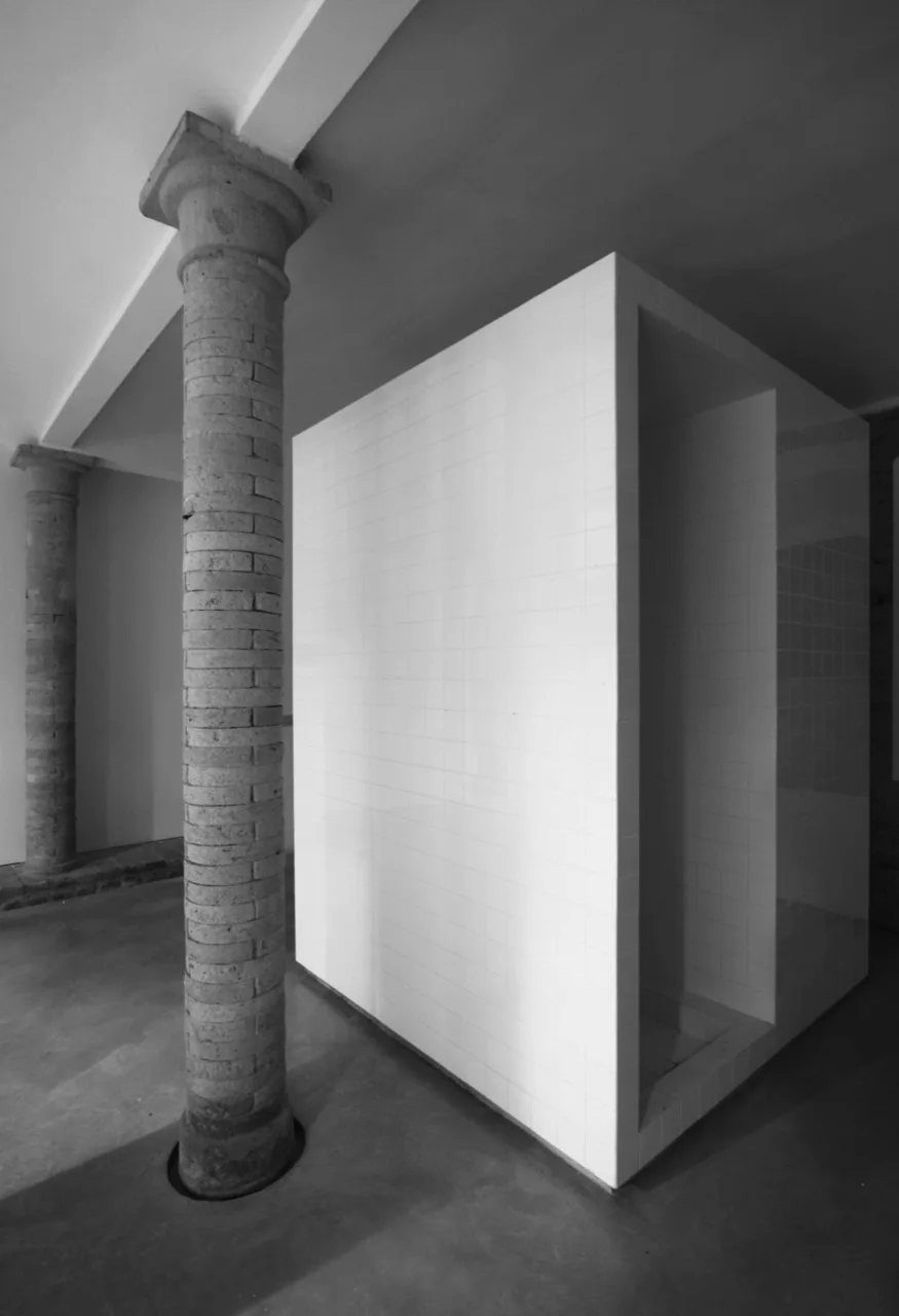 A white rectangular tiled structure stands on a smooth concrete floor next to two tall, textured stone columns.