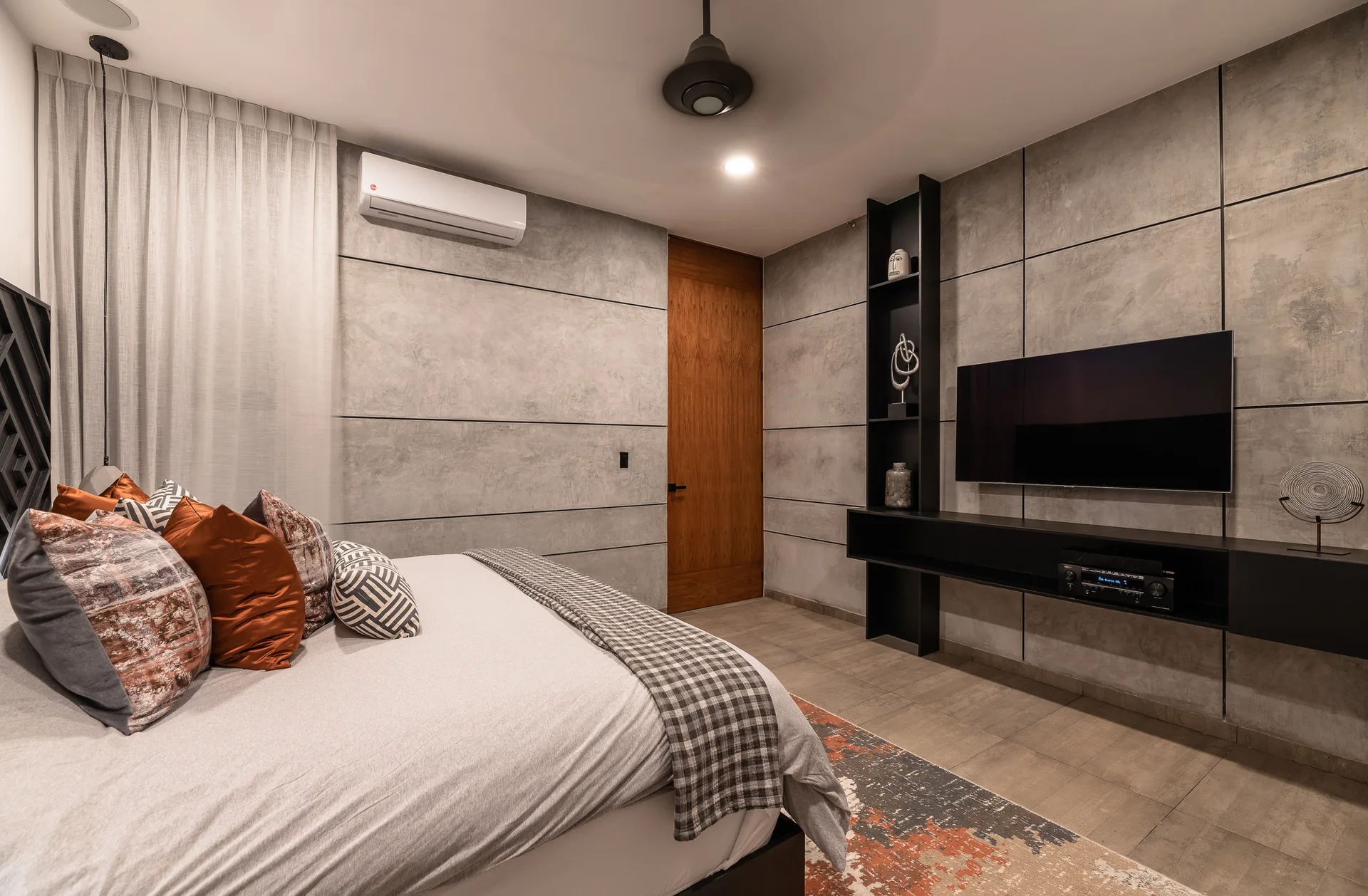 A modern bedroom with textured concrete wall panels, a bed featuring rust-orange and gray pillows, a wall-mounted TV, and a black floating console.