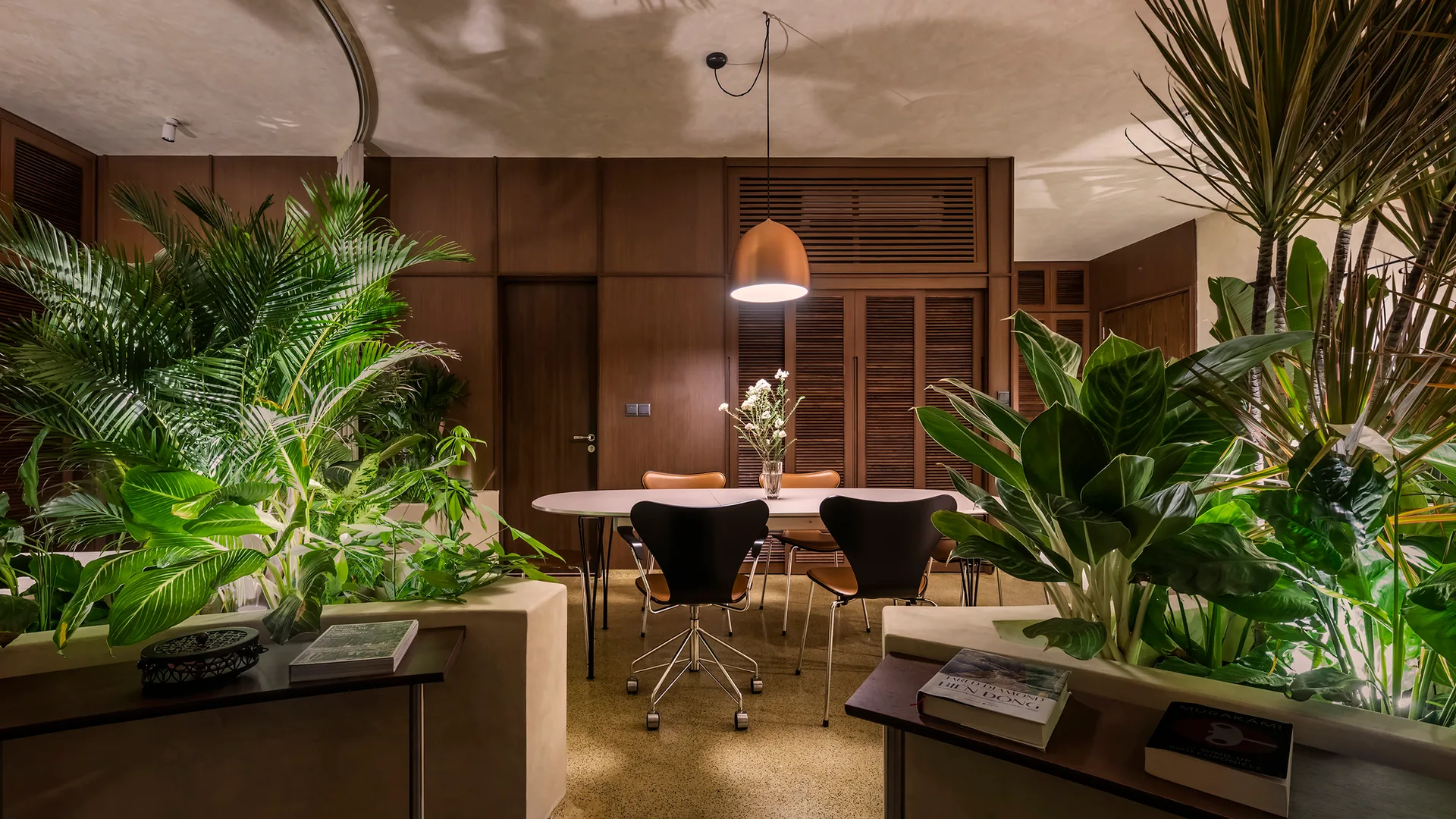 An oval dining table and chairs are framed by built-in planters with lush tropical foliage in a wood-paneled room.