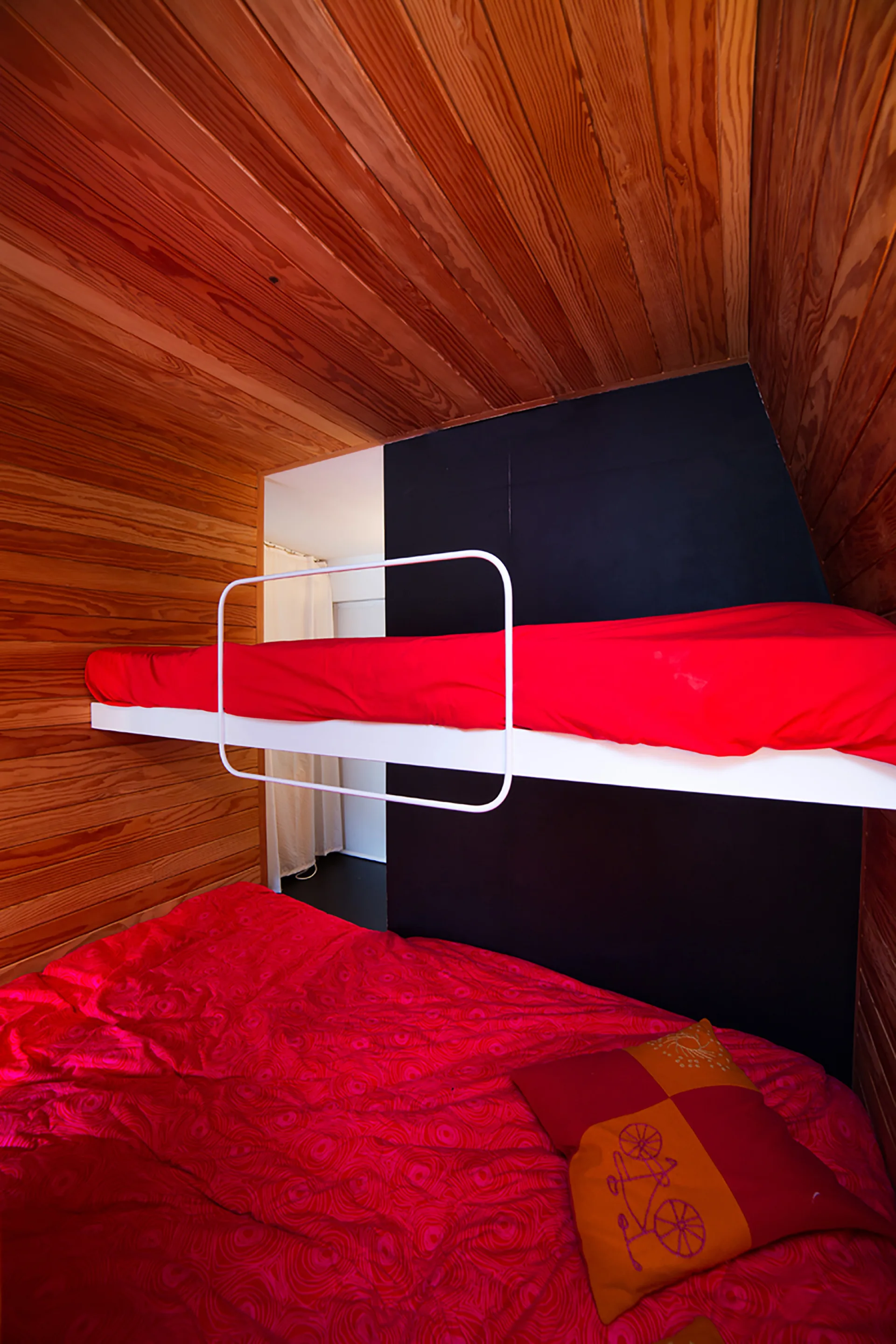 A compact bedroom features a white metal bunk bed with red bedding set against a black wall and wood paneled walls and ceiling.