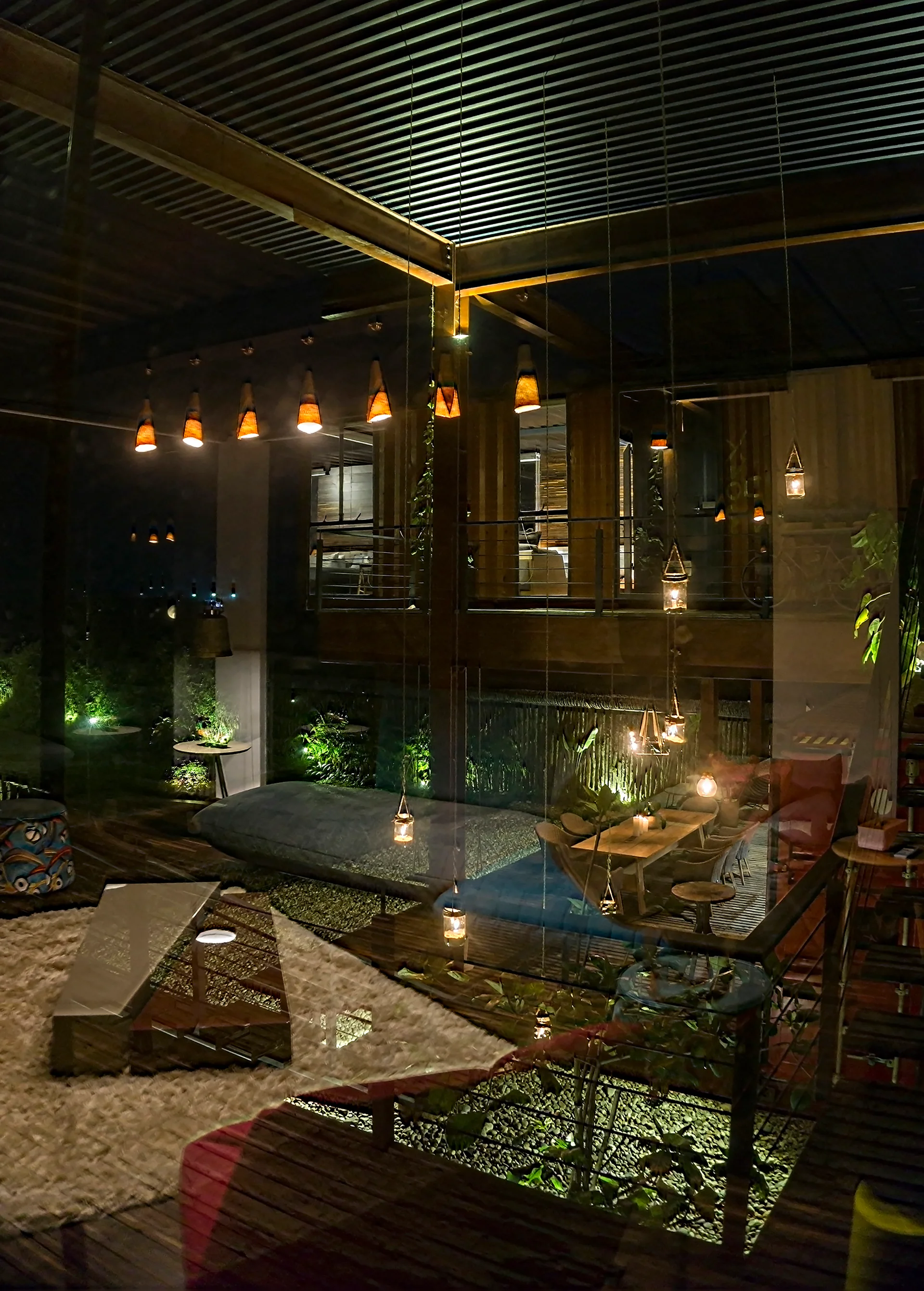 An outdoor courtyard at night featuring a wooden dining set, a low-profile lounge cushion on a rug, and suspended warm pendant lights.
