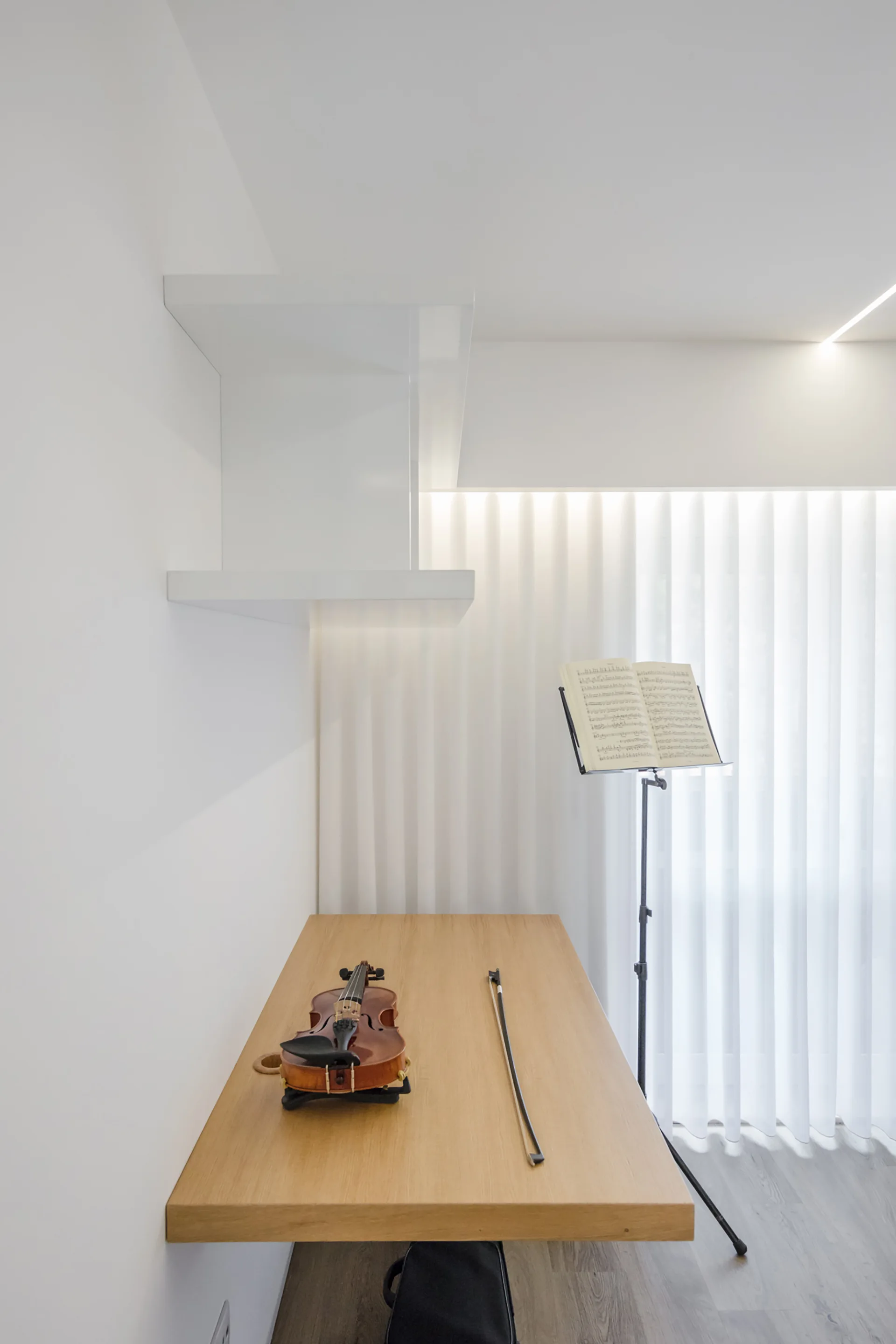 A minimalist music workspace features a natural wood desk holding a violin, positioned next to a black music stand in front of white ripple-fold curtains.