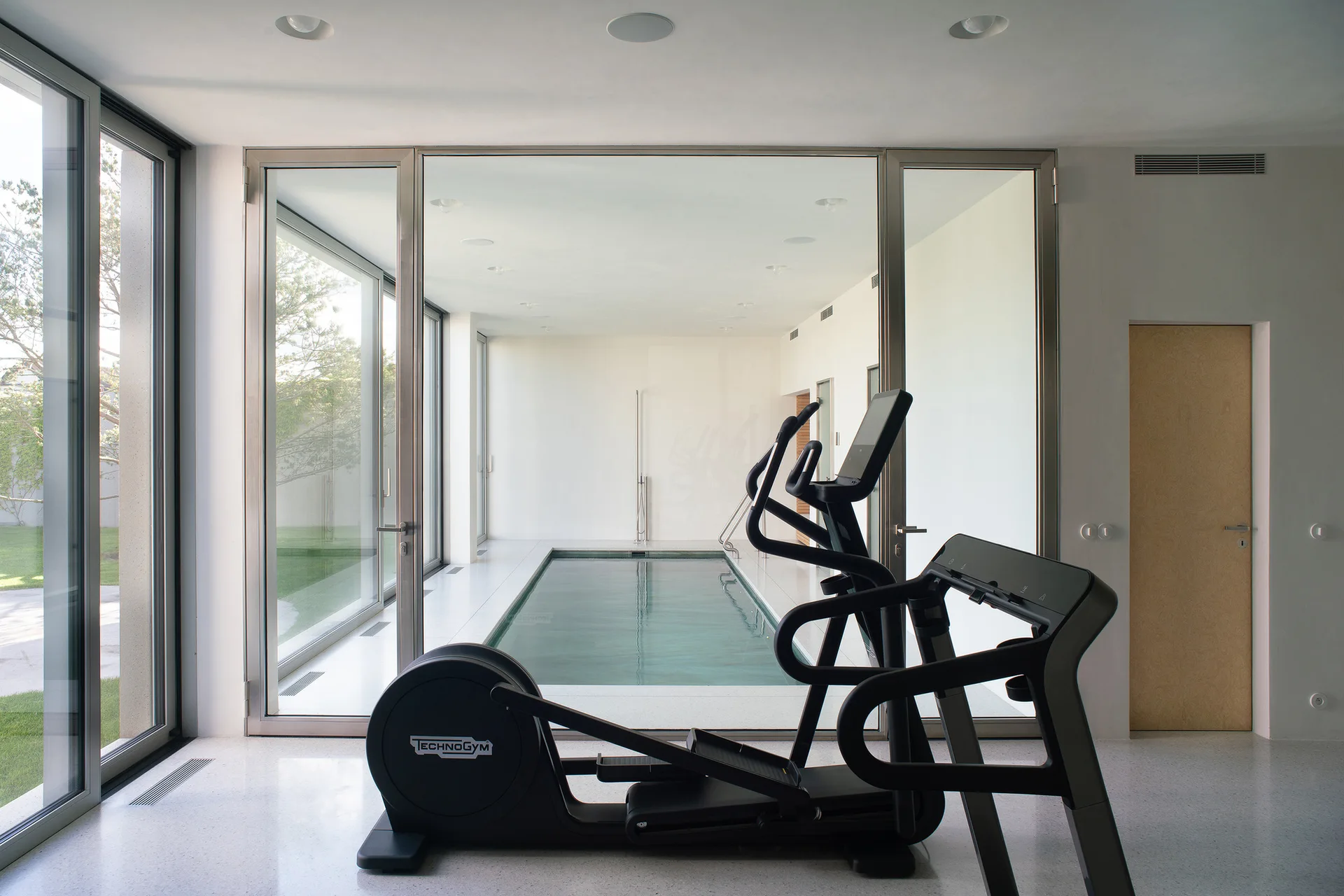 A black elliptical trainer sits on a light grey stone floor in front of an indoor lap pool enclosed by floor-to-ceiling glass walls.