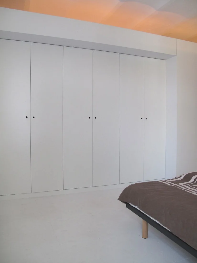 A minimalist bedroom featuring a large white floor-to-ceiling built-in wardrobe with small black door pulls next to a bed with brown covers.
