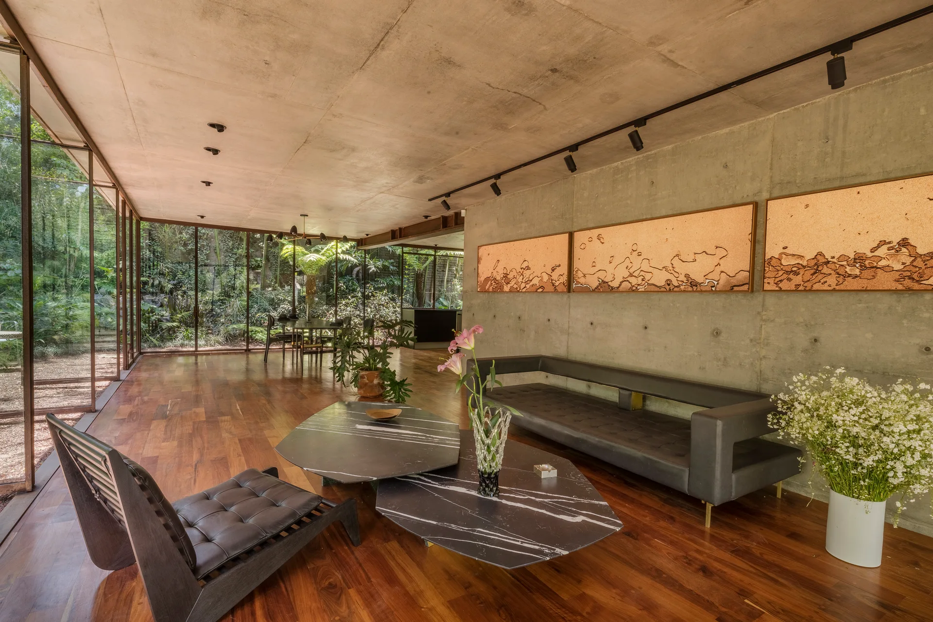 A modern living room features raw concrete walls, warm wood flooring, a low-profile sofa, and large glass windows overlooking a lush garden.