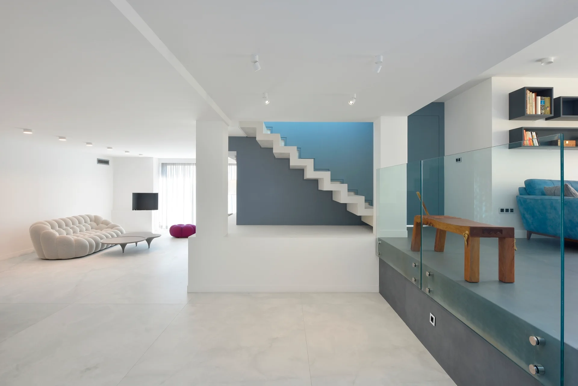 A minimalist split-level interior featuring a curved modular sofa, a white floating staircase against a blue wall, and an elevated glass-railed platform.