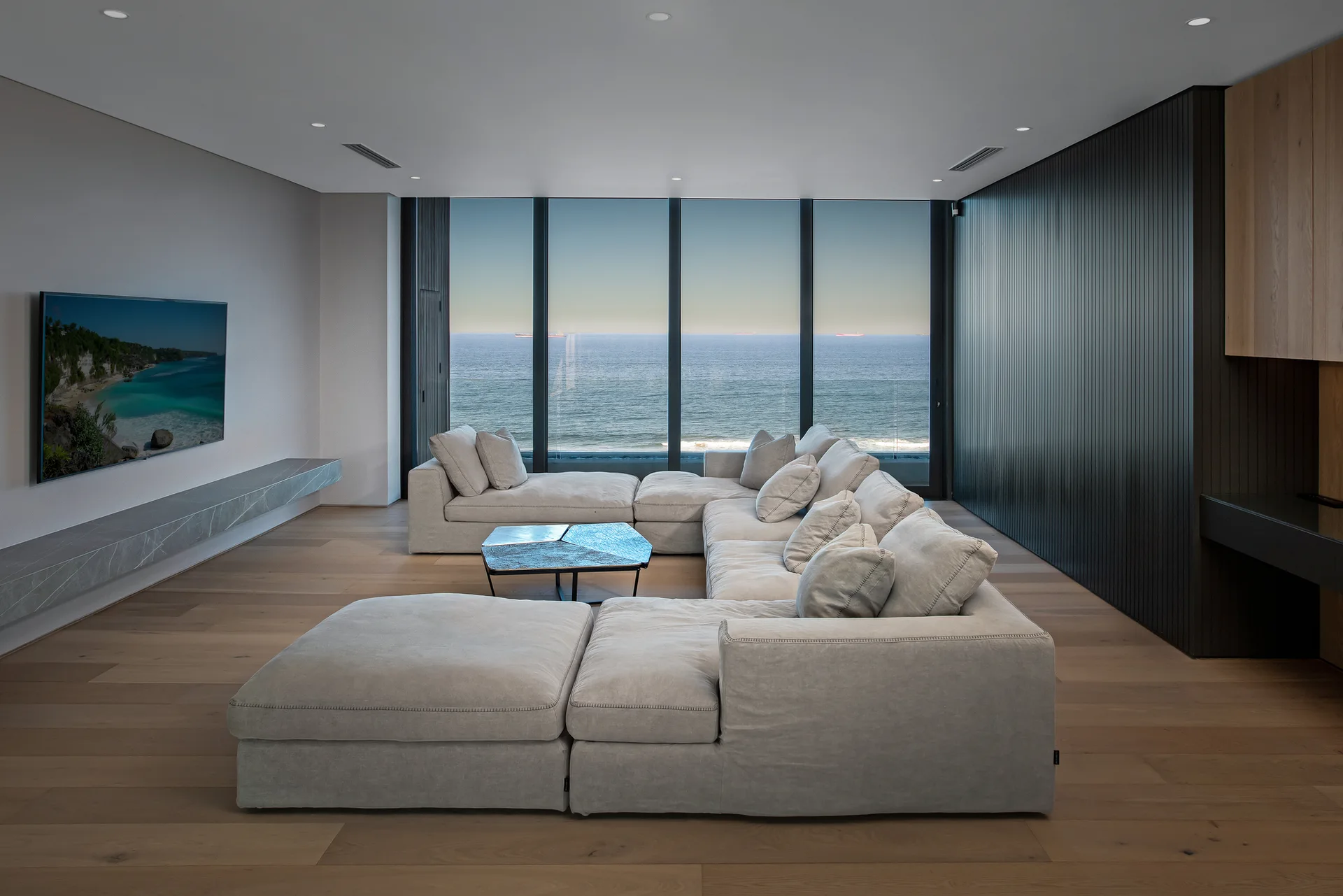 A spacious living room features a large light gray sectional, a dark slatted accent wall, and panoramic windows overlooking the ocean.