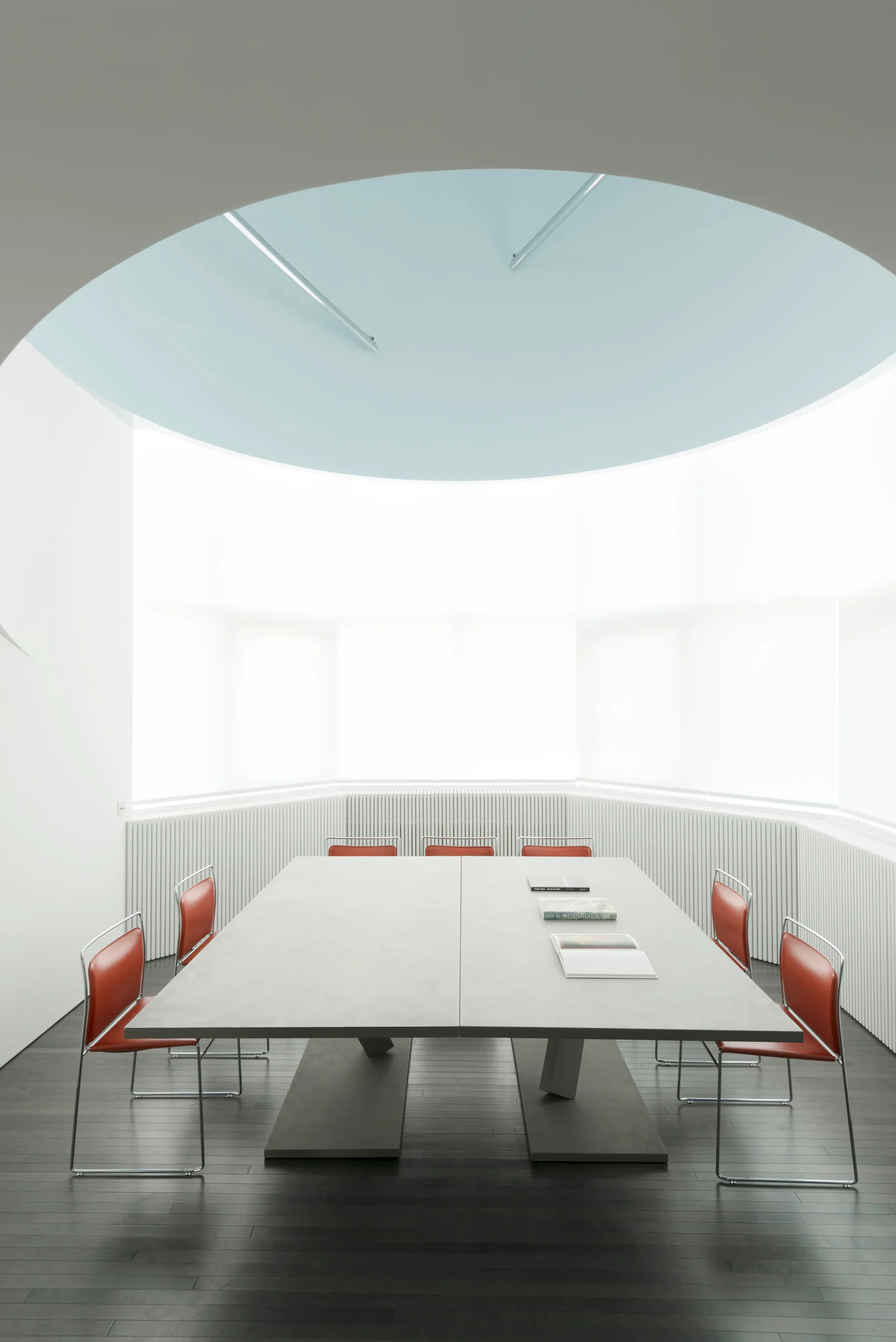 A minimalist room features a grey table surrounded by red chairs in front of a curved, slatted wall below a pale blue circular ceiling.