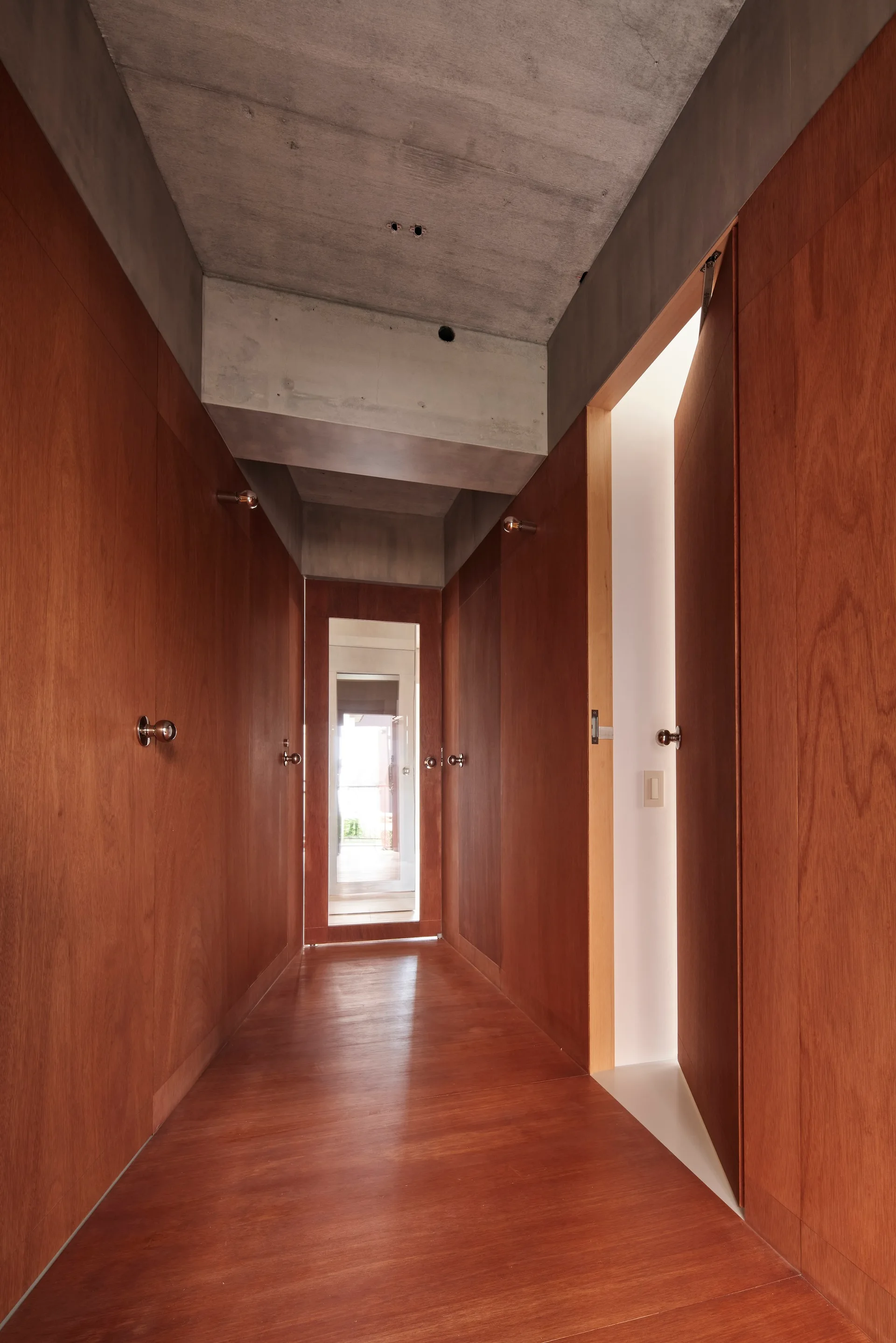 A narrow modern hallway featuring warm wood-paneled walls, matching wood floors, flush doors with round knobs, and an exposed concrete ceiling.