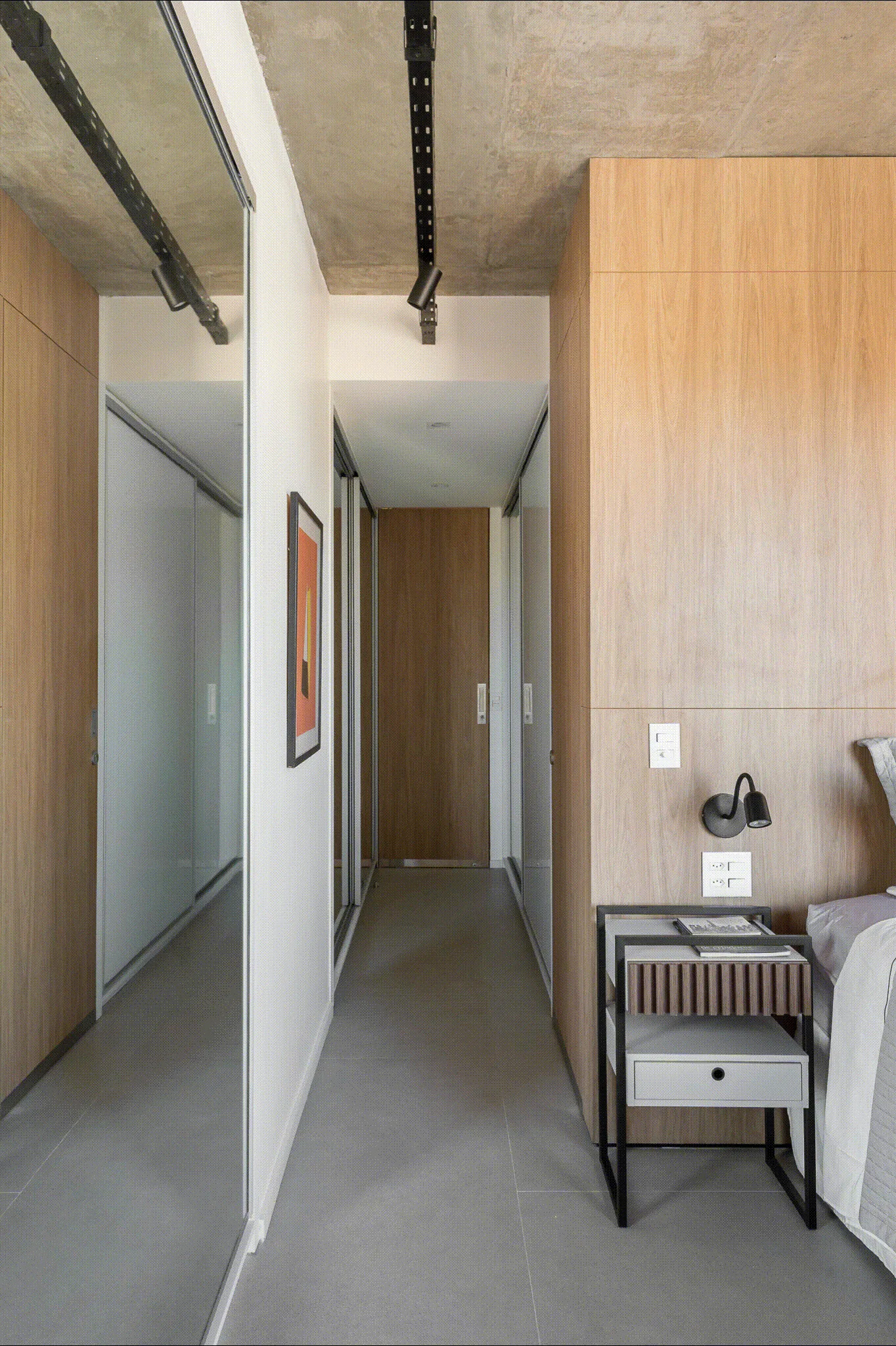 A modern bedroom entryway features a textured concrete ceiling, built-in light wood closets, a full-length mirrored door, and a sleek grey floor.