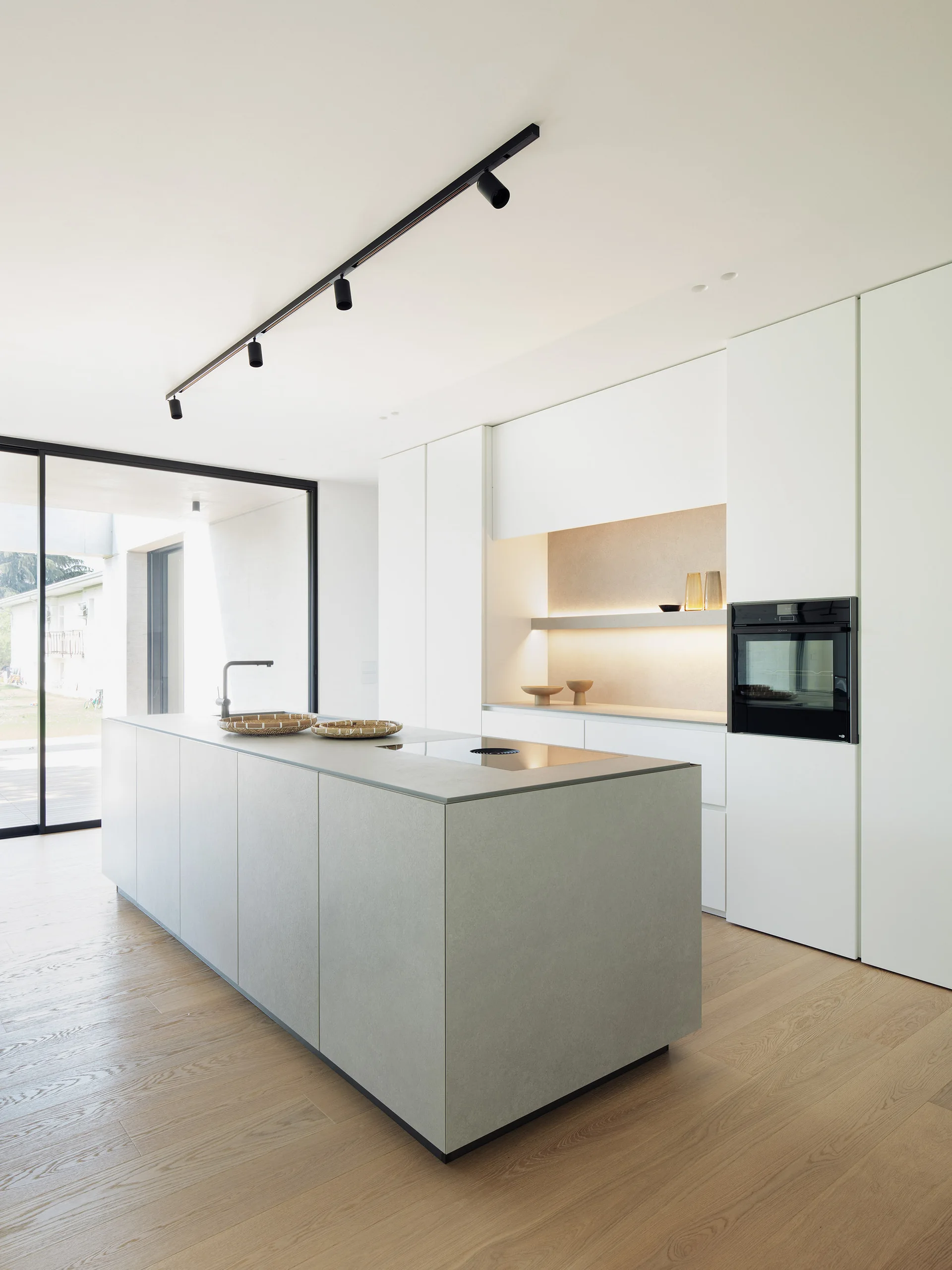 A minimalist modern kitchen featuring a light grey matte island, tall white handleless cabinets with an integrated shelf, and light wood flooring.