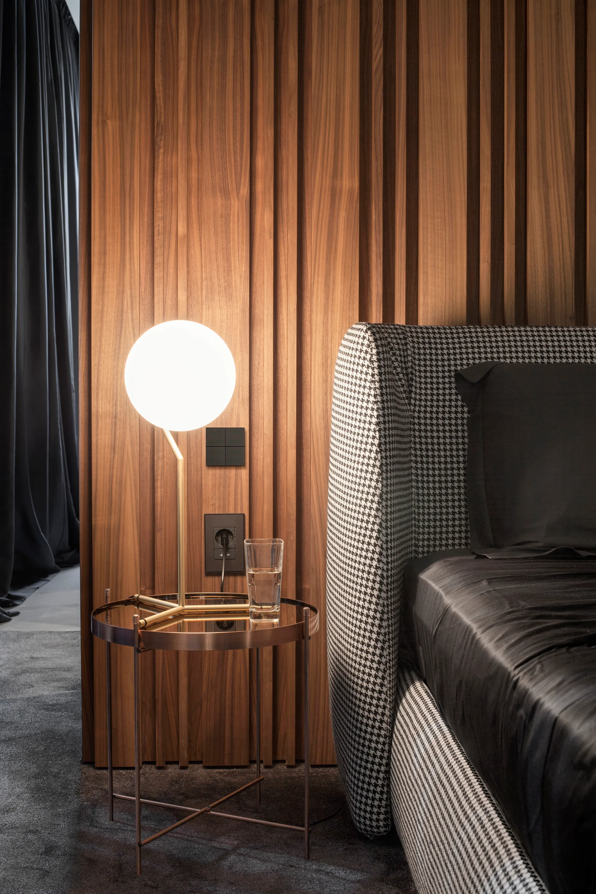 A bedroom details a houndstooth headboard, a round glass-topped metal nightstand with a lit globe lamp, and a vertically slatted wood accent wall.