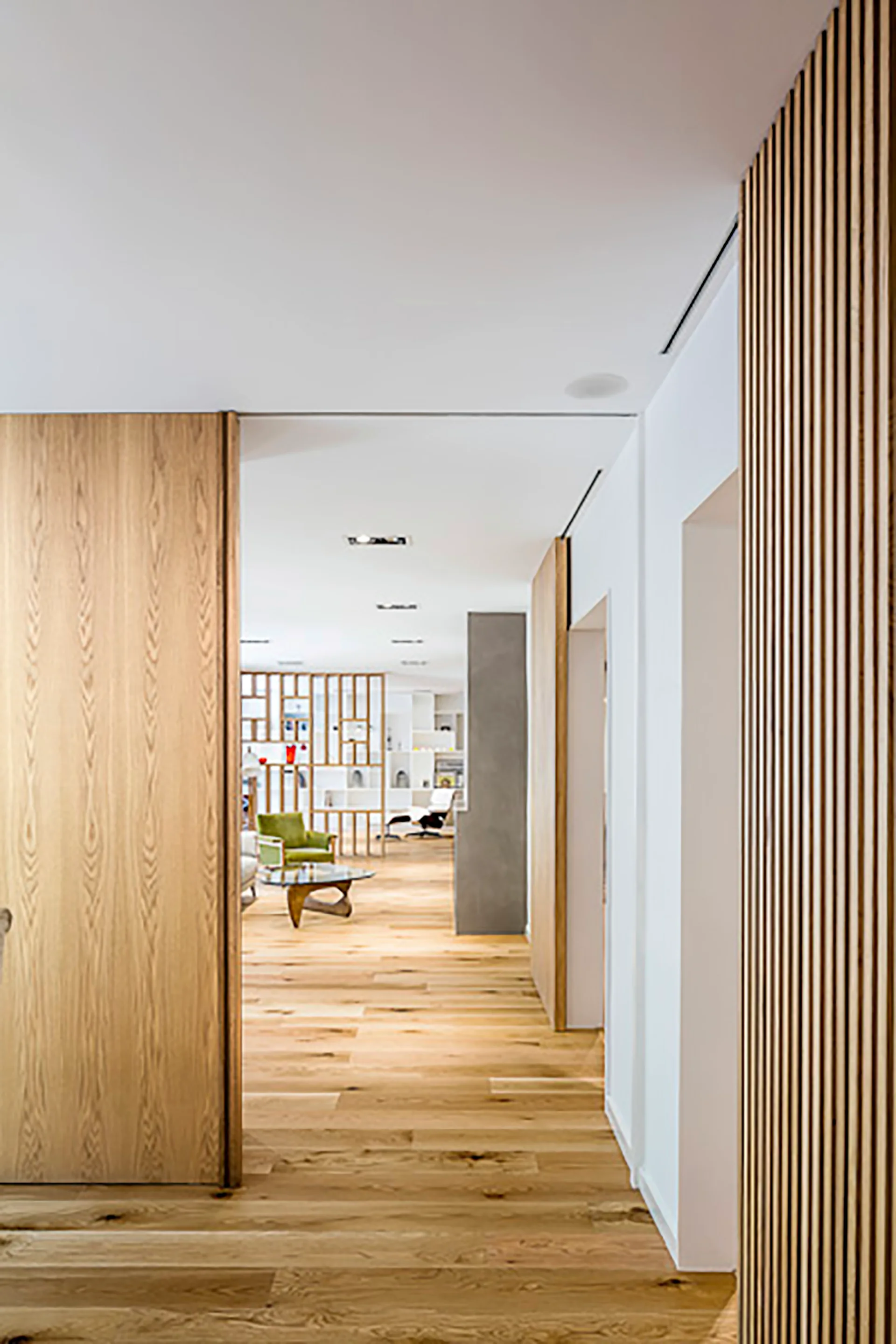 A modern hallway features a light wood pivot door, a vertical wood slat wall, and hardwood flooring leading into a living room with a green armchair.
