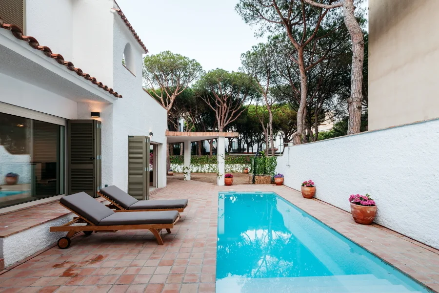 A rectangular swimming pool is flanked by a terracotta tile patio, two wooden lounge chairs, a white stucco wall, and tall pine trees.