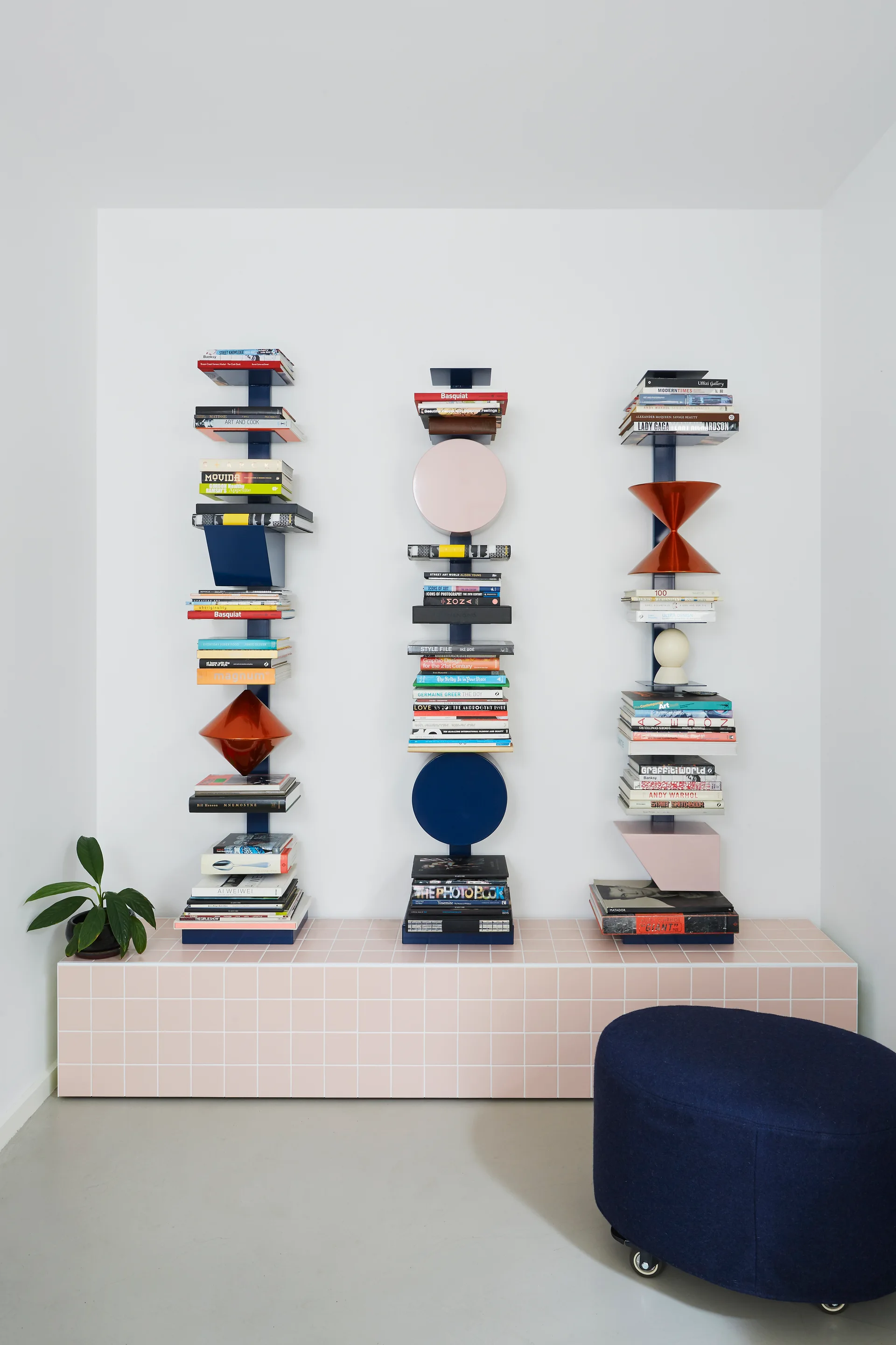 Three vertical navy blue spine bookshelves with books and colorful geometric shapes sit atop a long pastel pink tiled bench next to a blue ottoman.