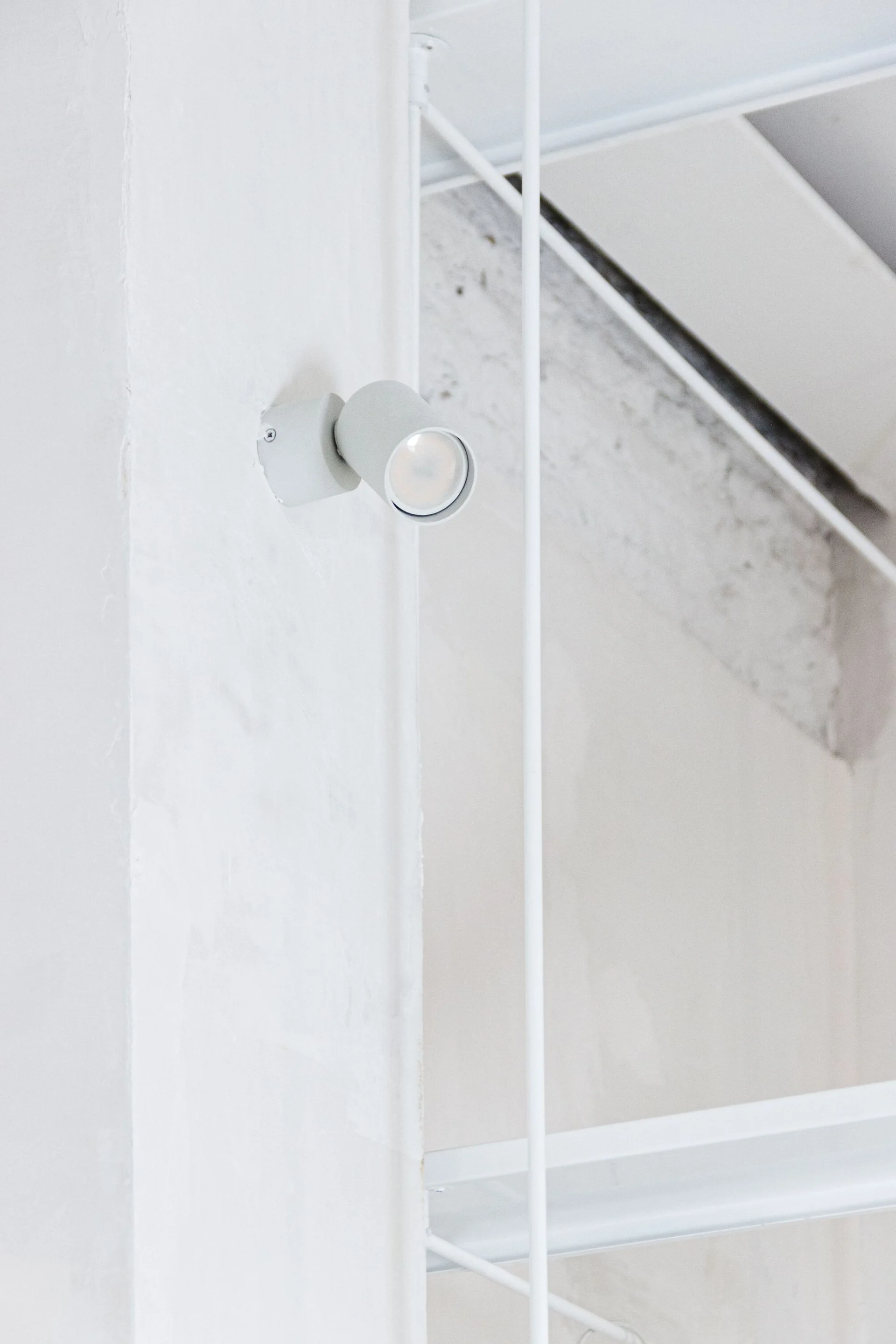 A small cylindrical white spotlight is mounted on a textured white wall next to slender white metal shelving supports.