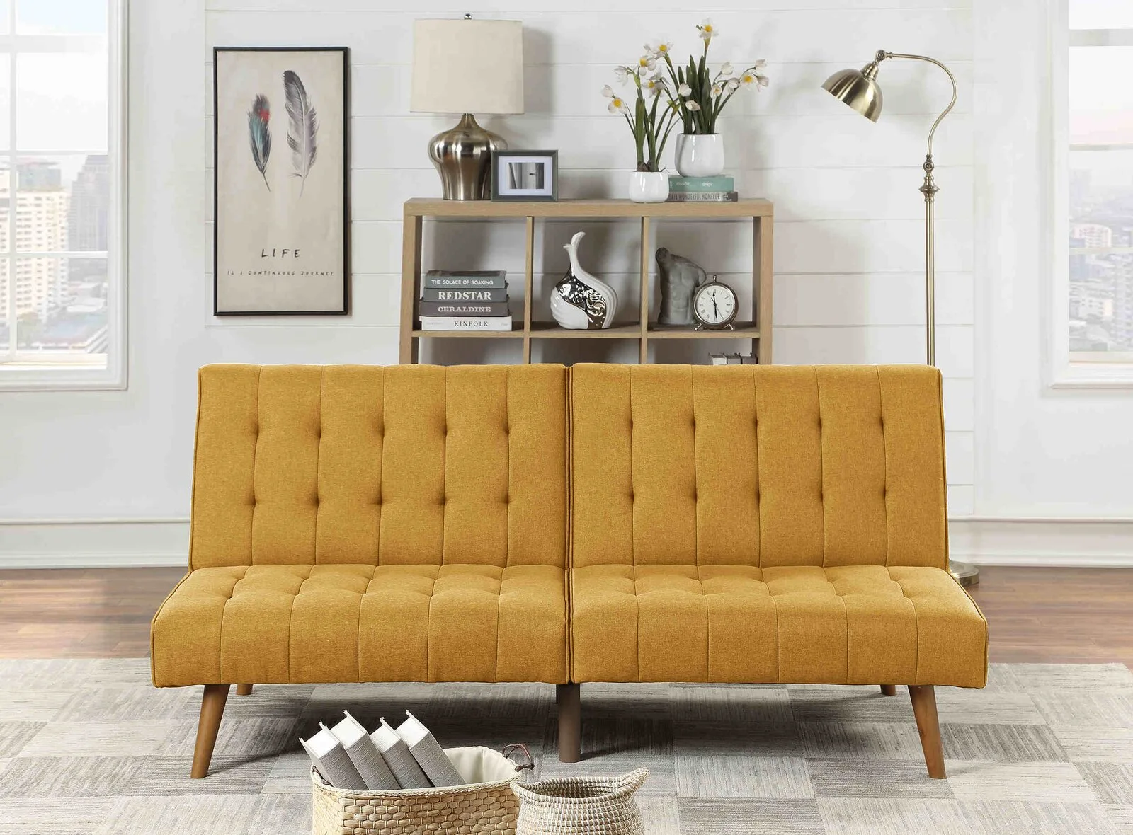 A mustard yellow tufted sofa sits on a textured rug in front of a light wood bookshelf, a metallic floor lamp, and a white shiplap wall.