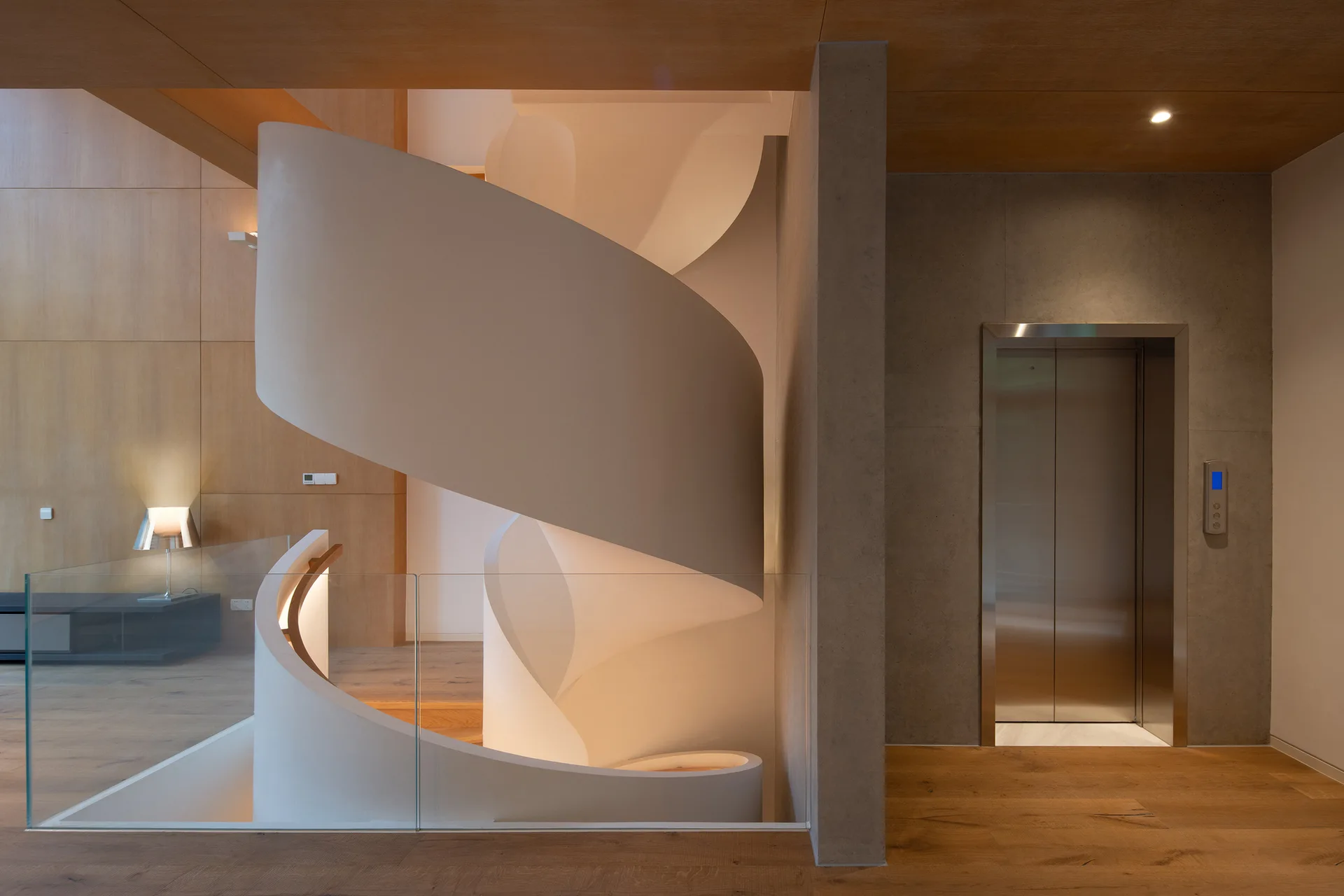 A modern sculptural white spiral staircase stands next to a stainless steel elevator surrounded by warm wood walls and flooring.