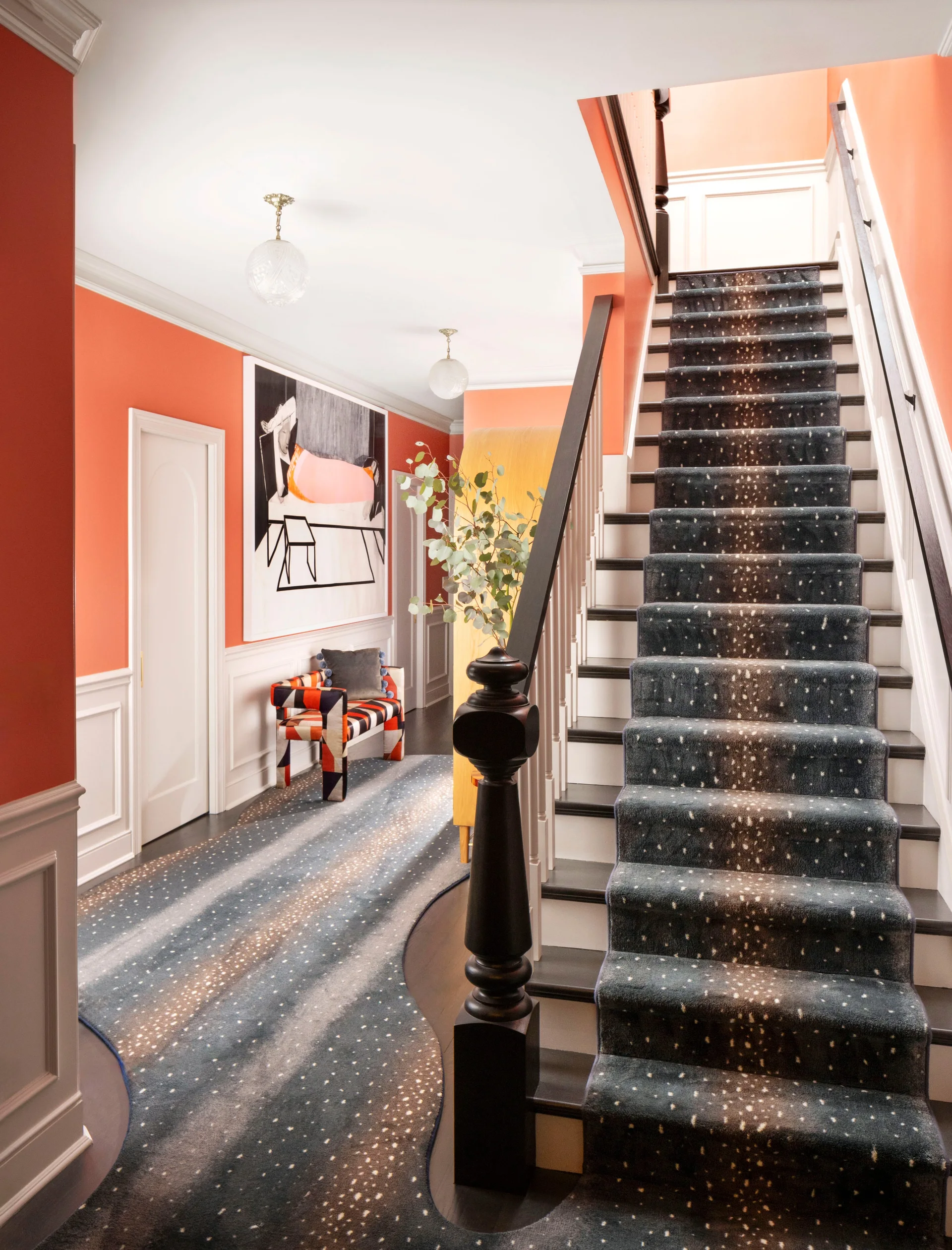 An entryway featuring bright coral walls, white wainscoting, and a dark wood staircase covered in a speckled carpet runner.