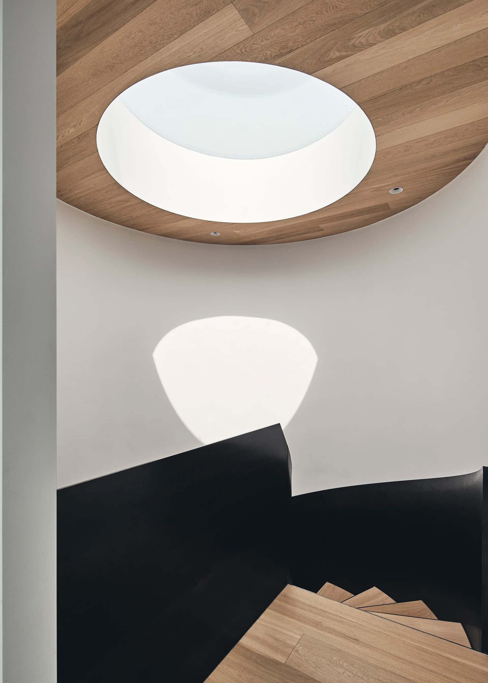 Minimalist spiral staircase with wood treads and a curved black railing below a circular skylight framed in wood planks.