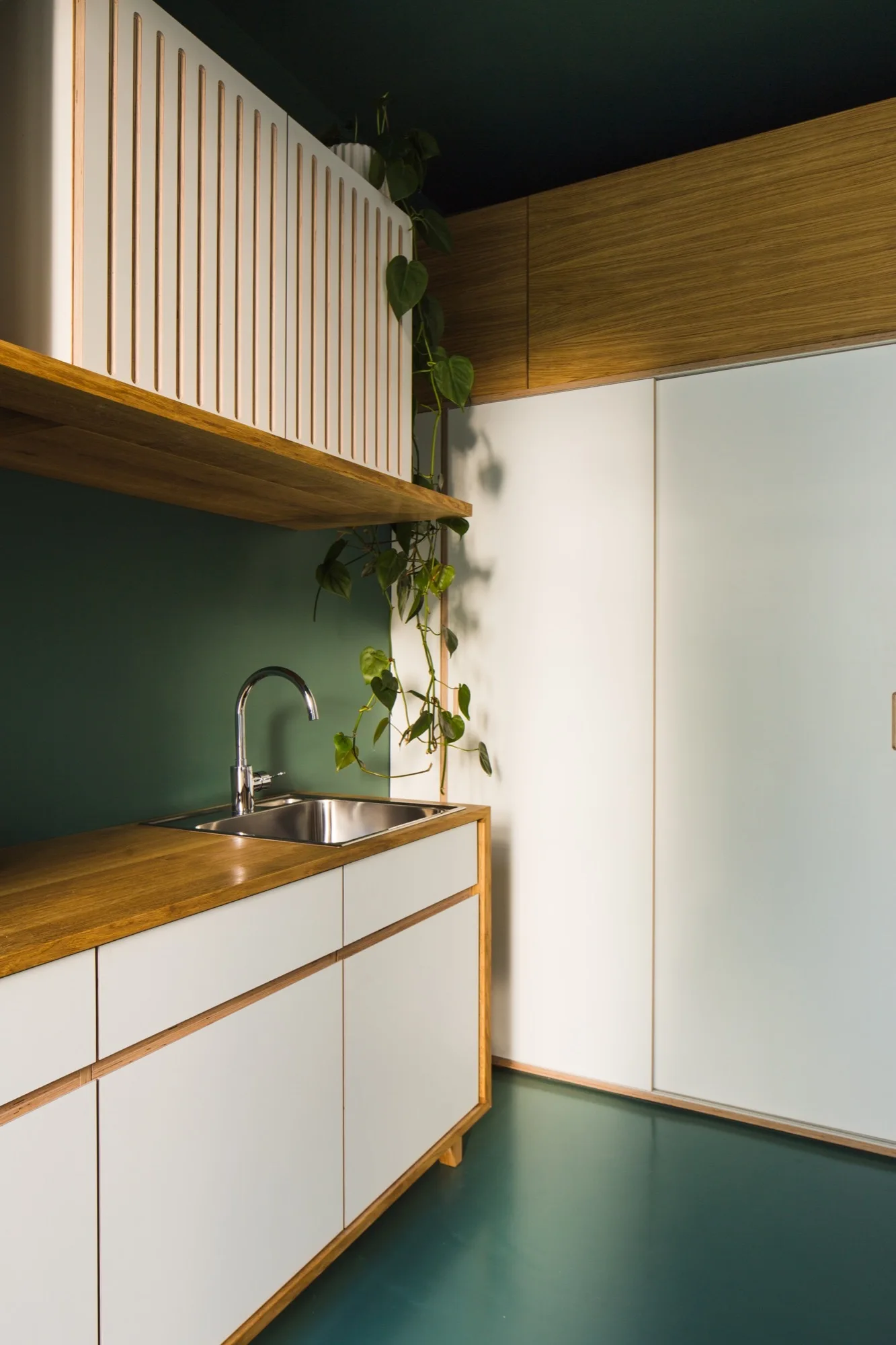 Modern kitchen corner featuring white cabinets with wood trim, a dark green backsplash, a small sink, and a vertically slatted upper cabinet.