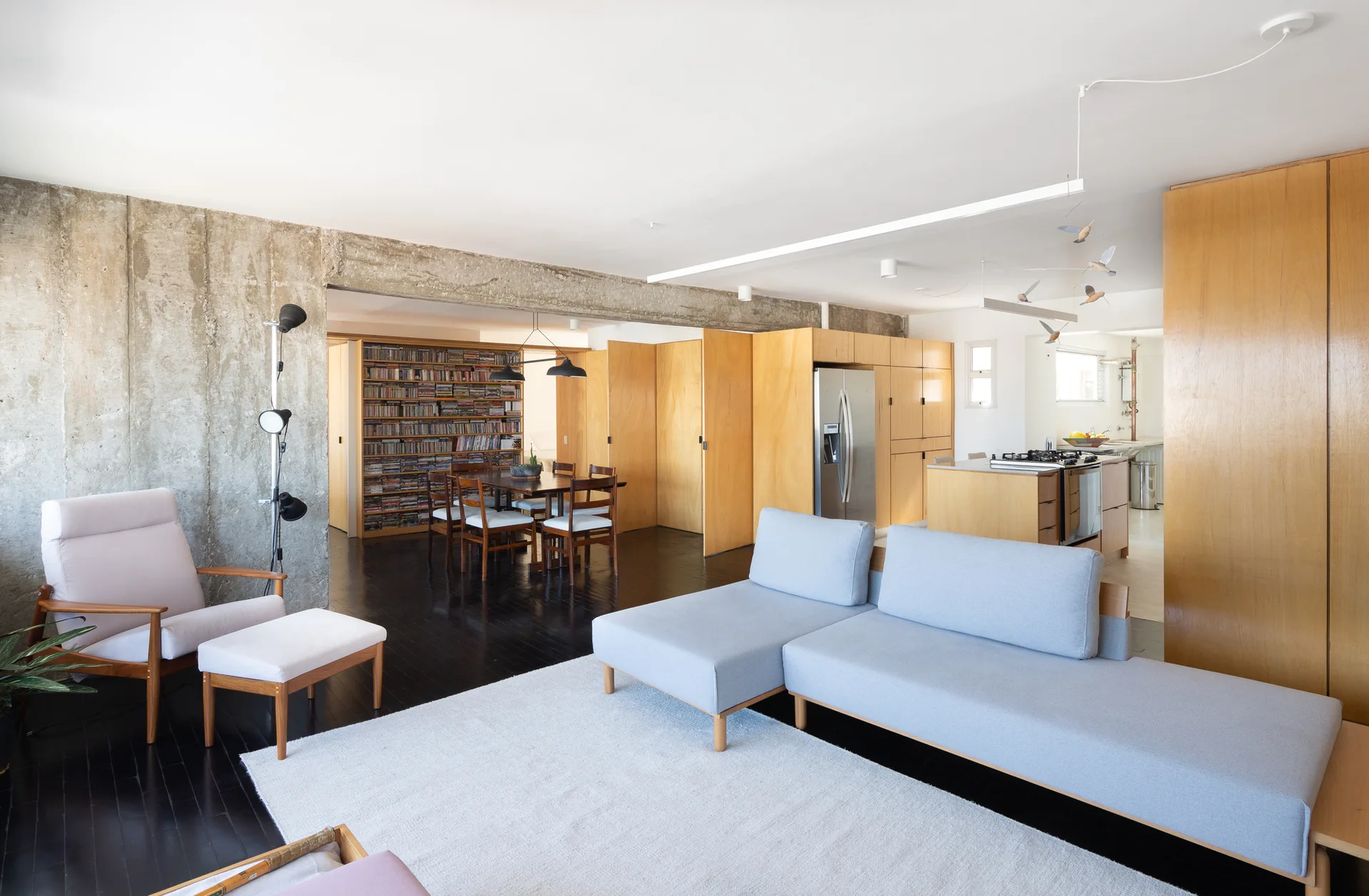 An open-concept living room features a light gray sectional sofa, a raw concrete beam overhead, and built-in light wood cabinetry.