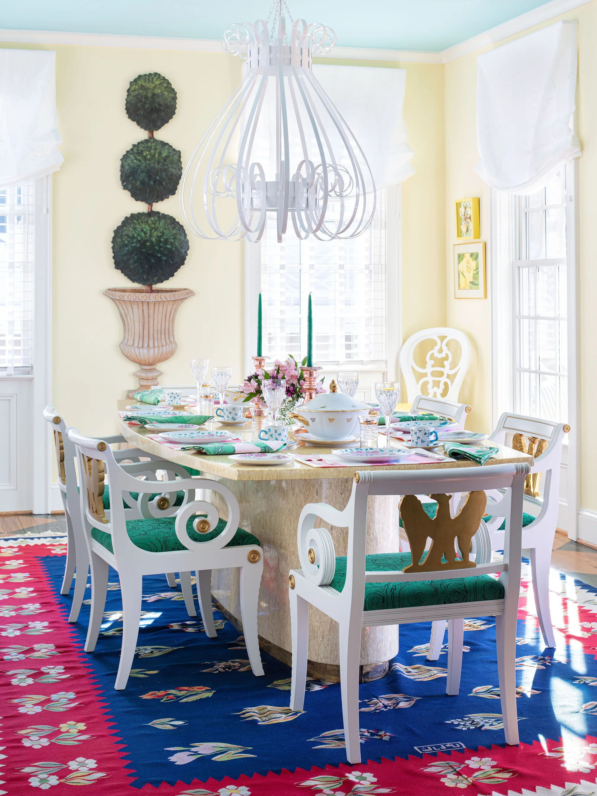 A yellow dining room features a stone-topped table set with dinnerware, white chairs with green cushions, and a white scrollwork chandelier.