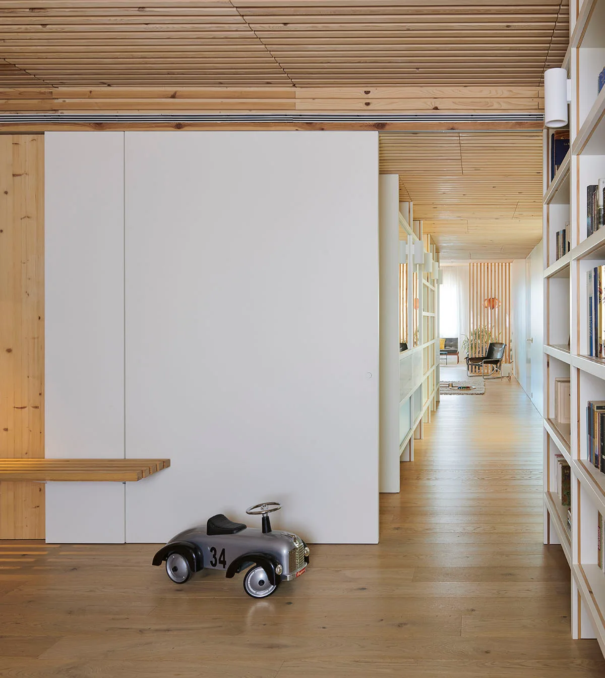 A minimalist hallway features light wood plank floors, a slatted wood ceiling, built-in bookshelves, and a large white sliding wall panel.