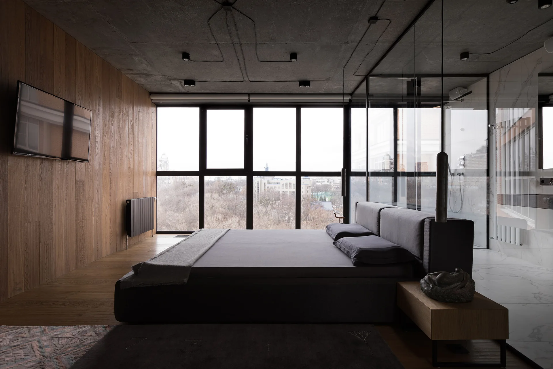 A minimalist bedroom featuring a grey platform bed, an exposed concrete ceiling, a wood-paneled wall, and a glass-partitioned bathroom.