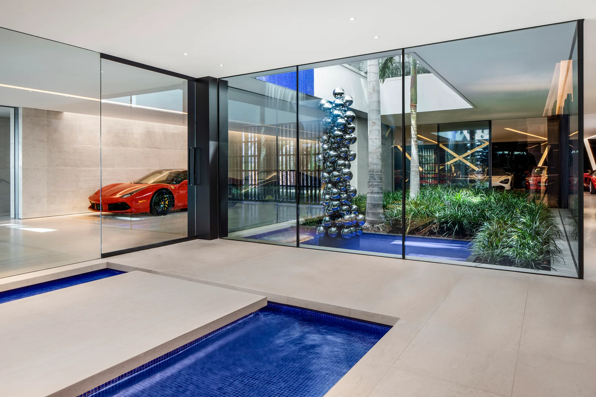 A minimalist home interior with beige tiled floors, floor-to-ceiling glass walls, a planted central courtyard, and a blue mosaic tile water feature.