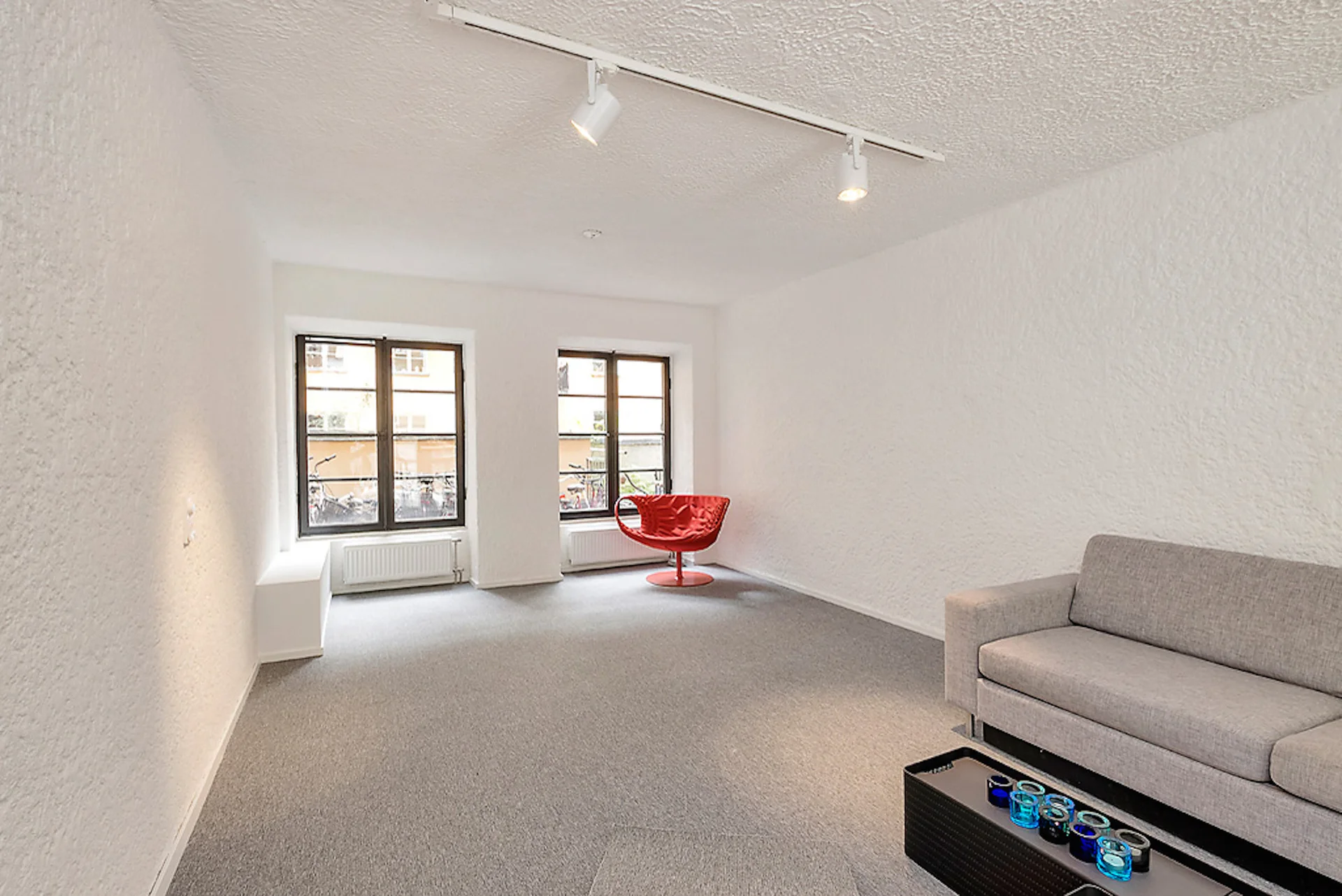 A minimalist living room features white textured stucco walls, a grey carpet, a modern grey sofa, and a vibrant red accent chair near two windows.