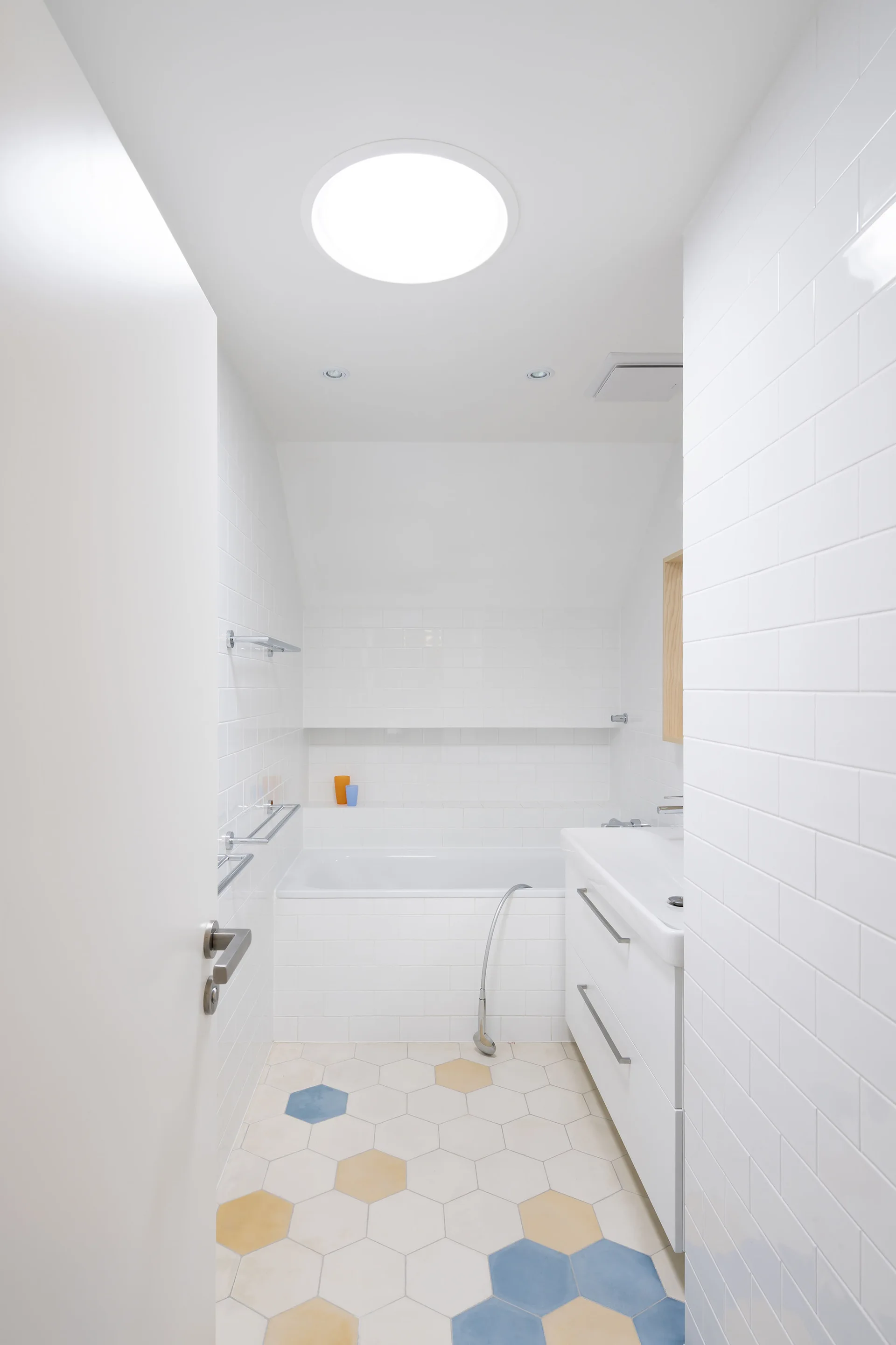 A modern white bathroom featuring glossy subway tile walls, a white bathtub, and patterned hexagonal floor tiles under a circular skylight.