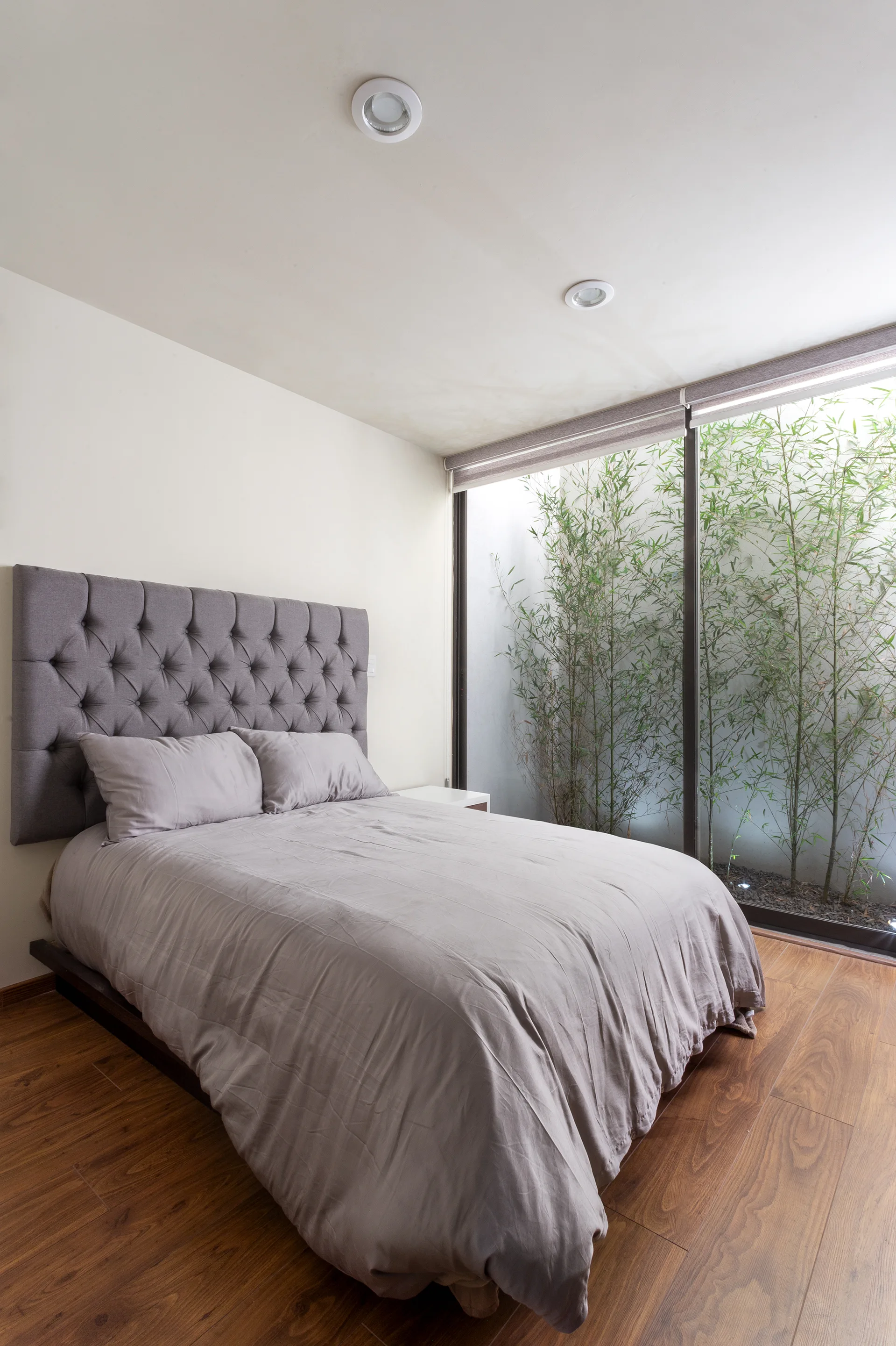 A modern bedroom featuring a bed with a tufted gray headboard next to a large sliding window looking out at bamboo plants.