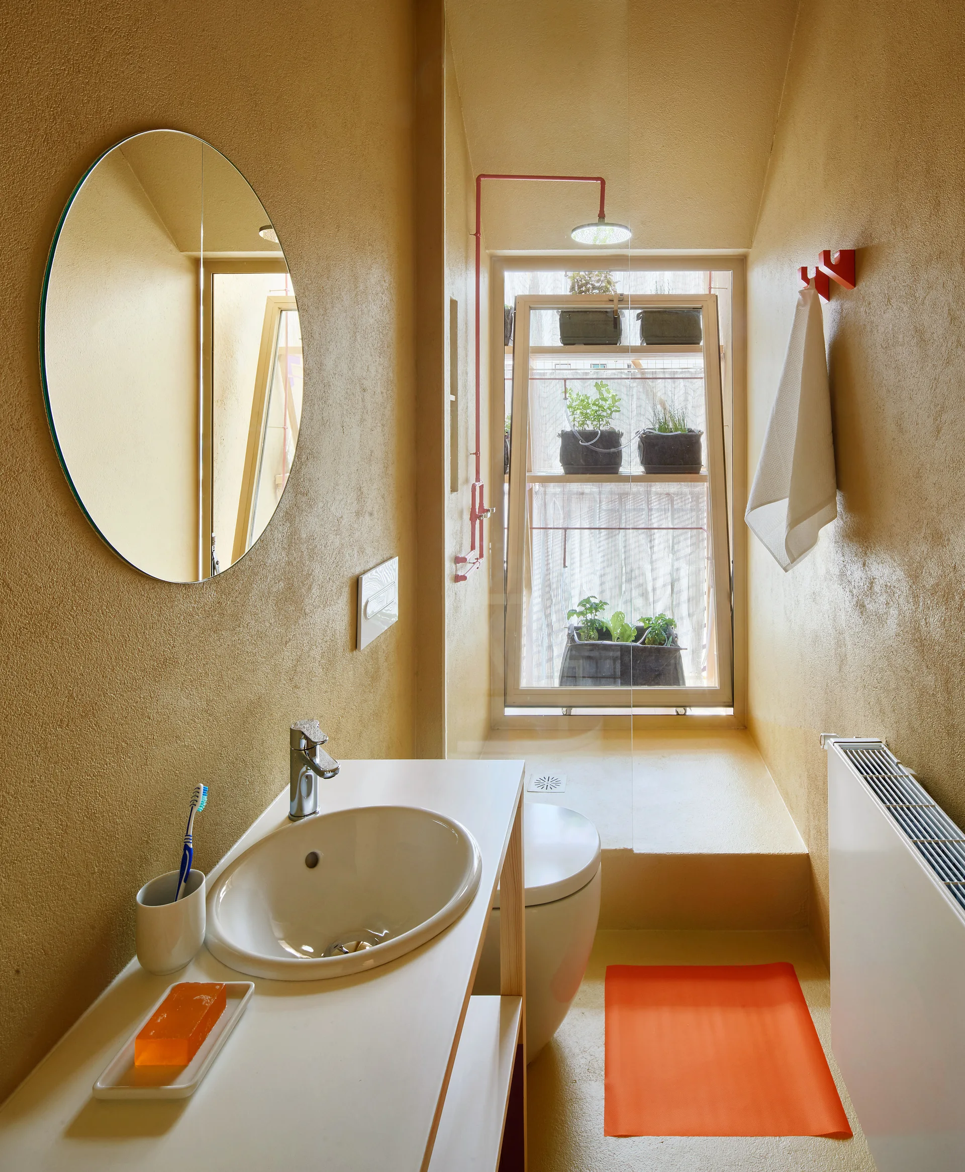 A narrow bathroom features textured ochre plaster walls, a round mirror, a white vanity sink, and bright red fixtures.
