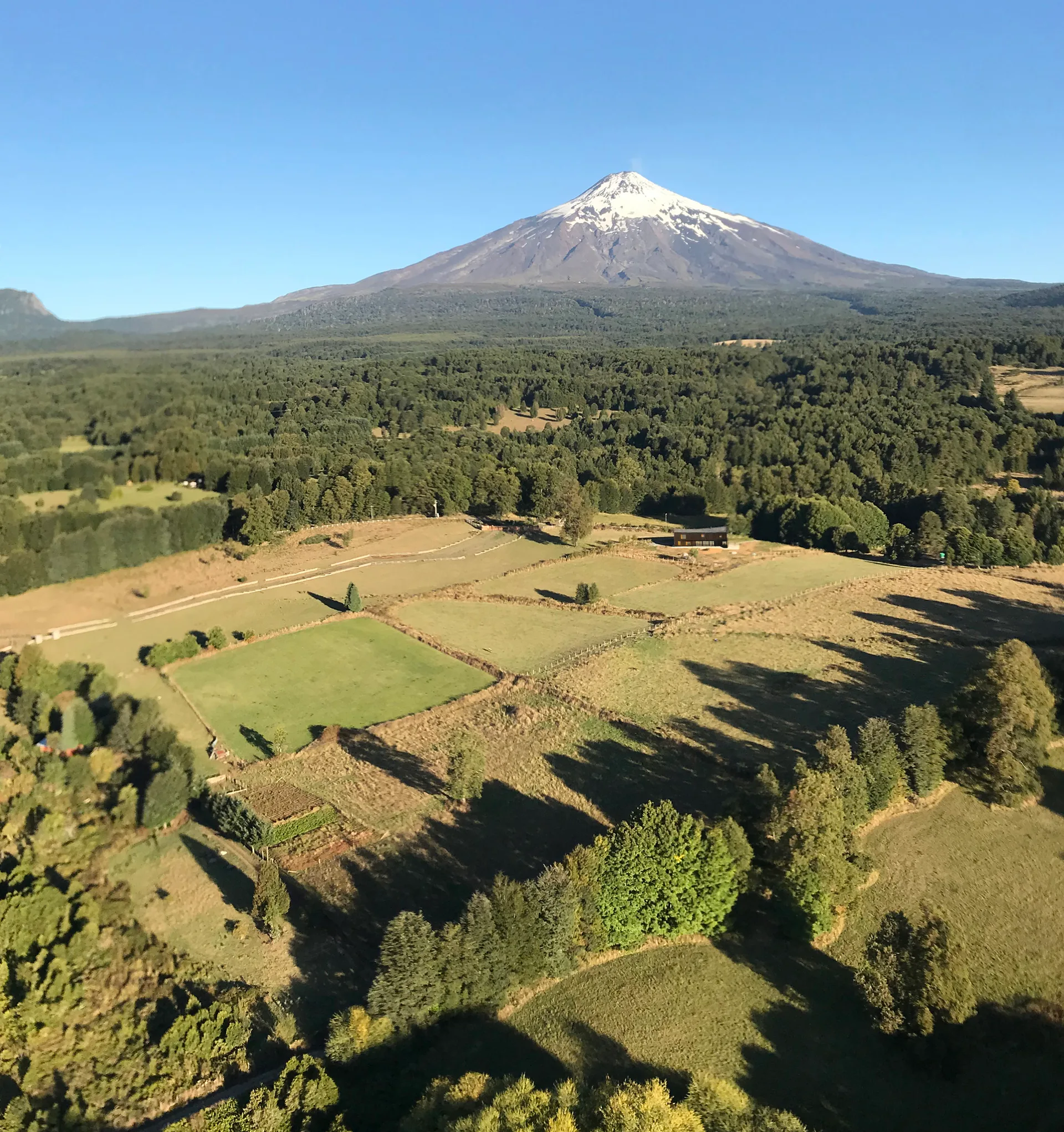 An aerial shot of a snow-capped volcanic mountain towering over green patchwork fields and lush forested hills under a clear blue sky.