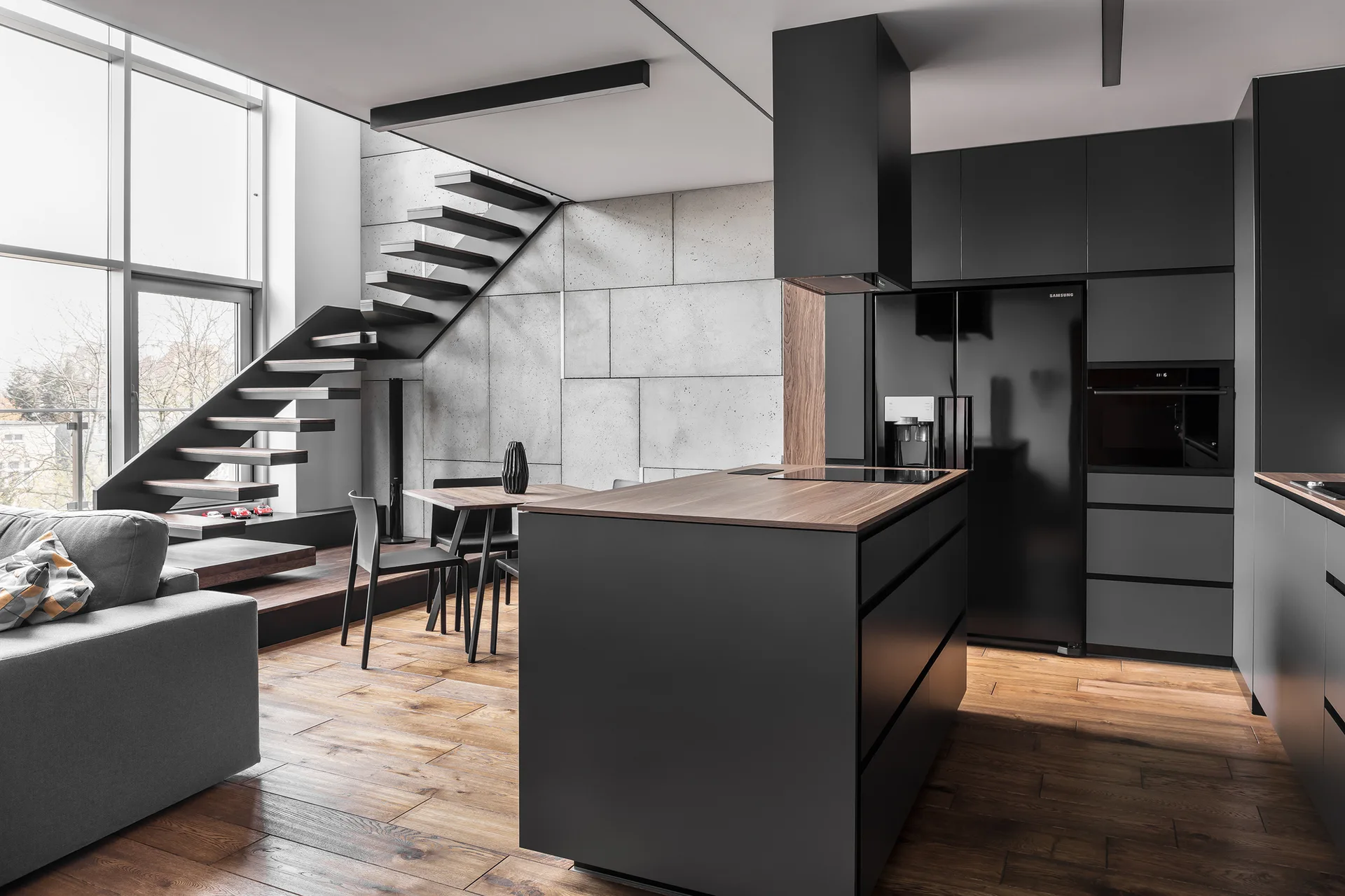 A modern minimalist kitchen with matte black cabinetry, a central island with a wood countertop, and a floating staircase against a concrete block wall.