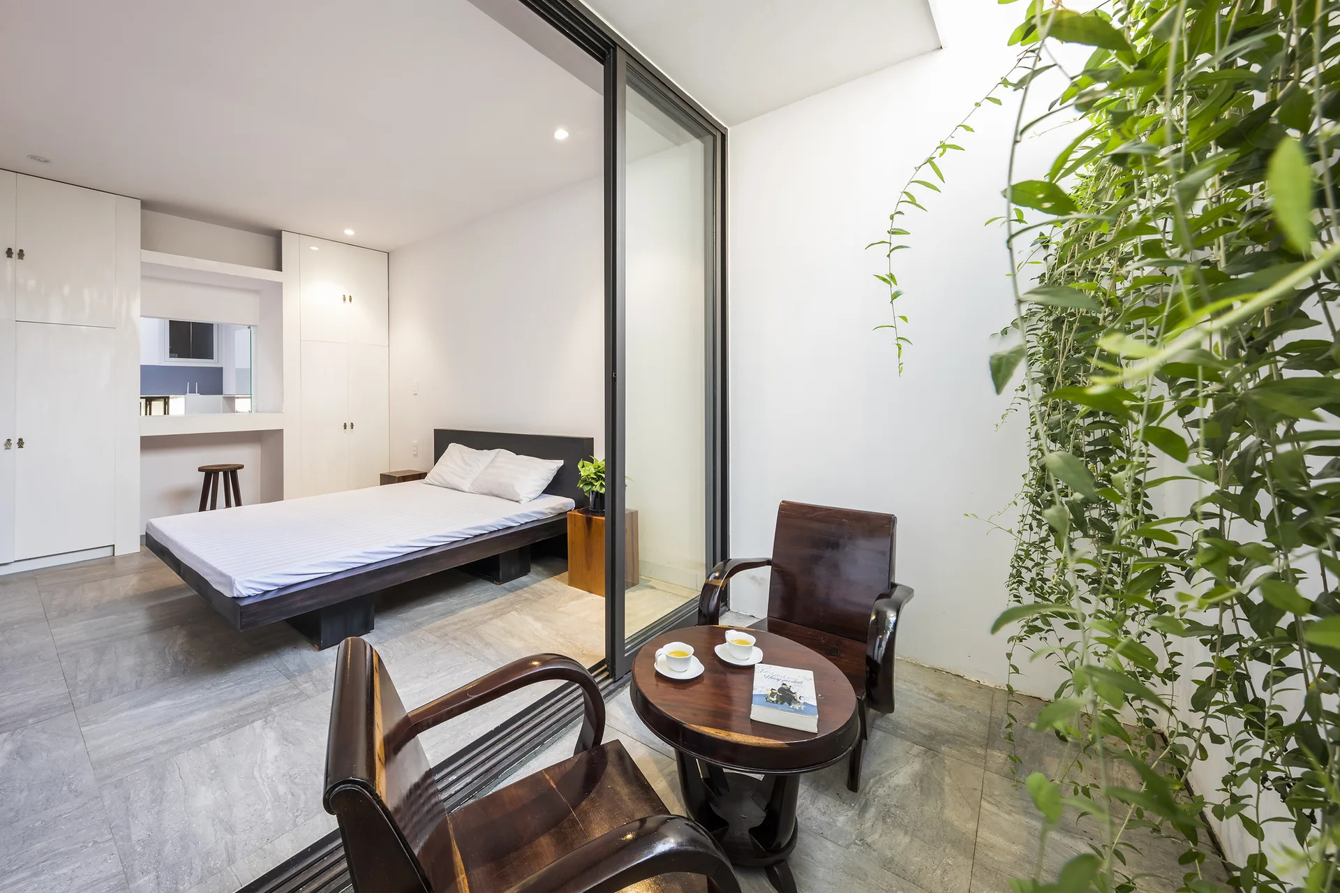 A minimalist bedroom with a low wood bed is separated from a tiled terrace with dark wood chairs by a large sliding glass door.
