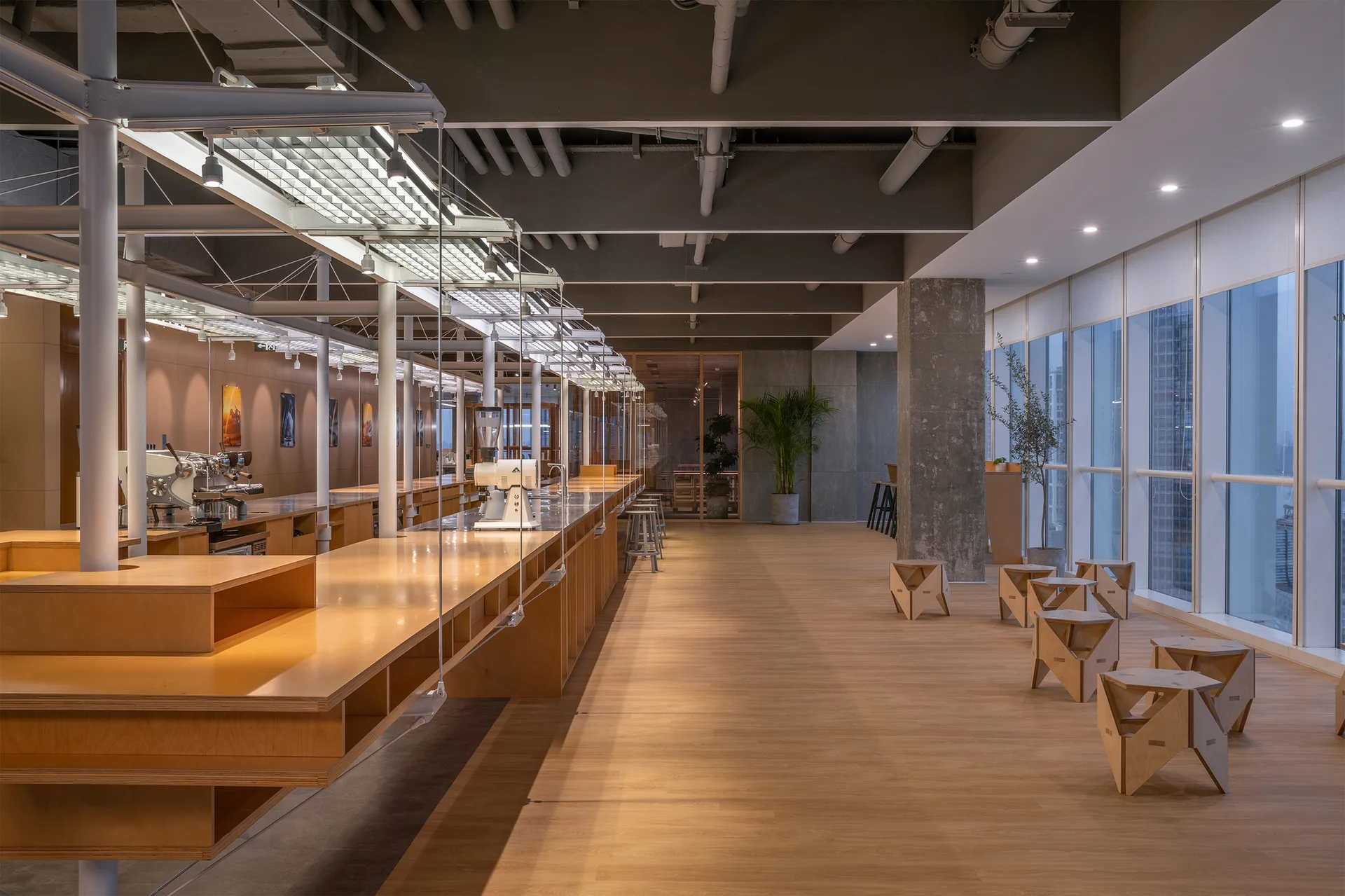 A spacious industrial interior features a long plywood counter with coffee equipment, geometric wood stools, and concrete columns.