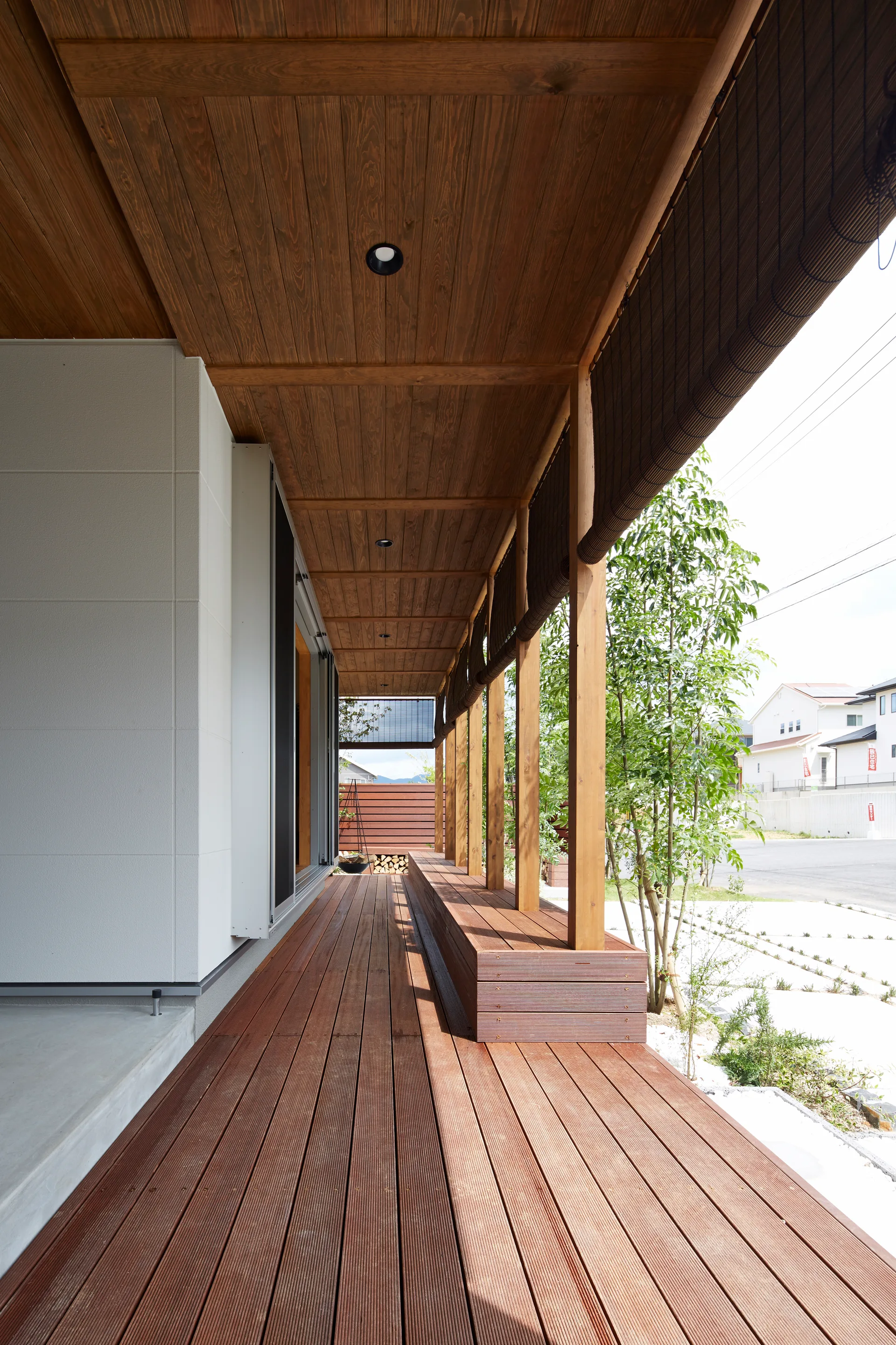 A long covered wooden porch features a built-in bench along the side, vertical support columns, and roll-up blinds hanging from the ceiling.