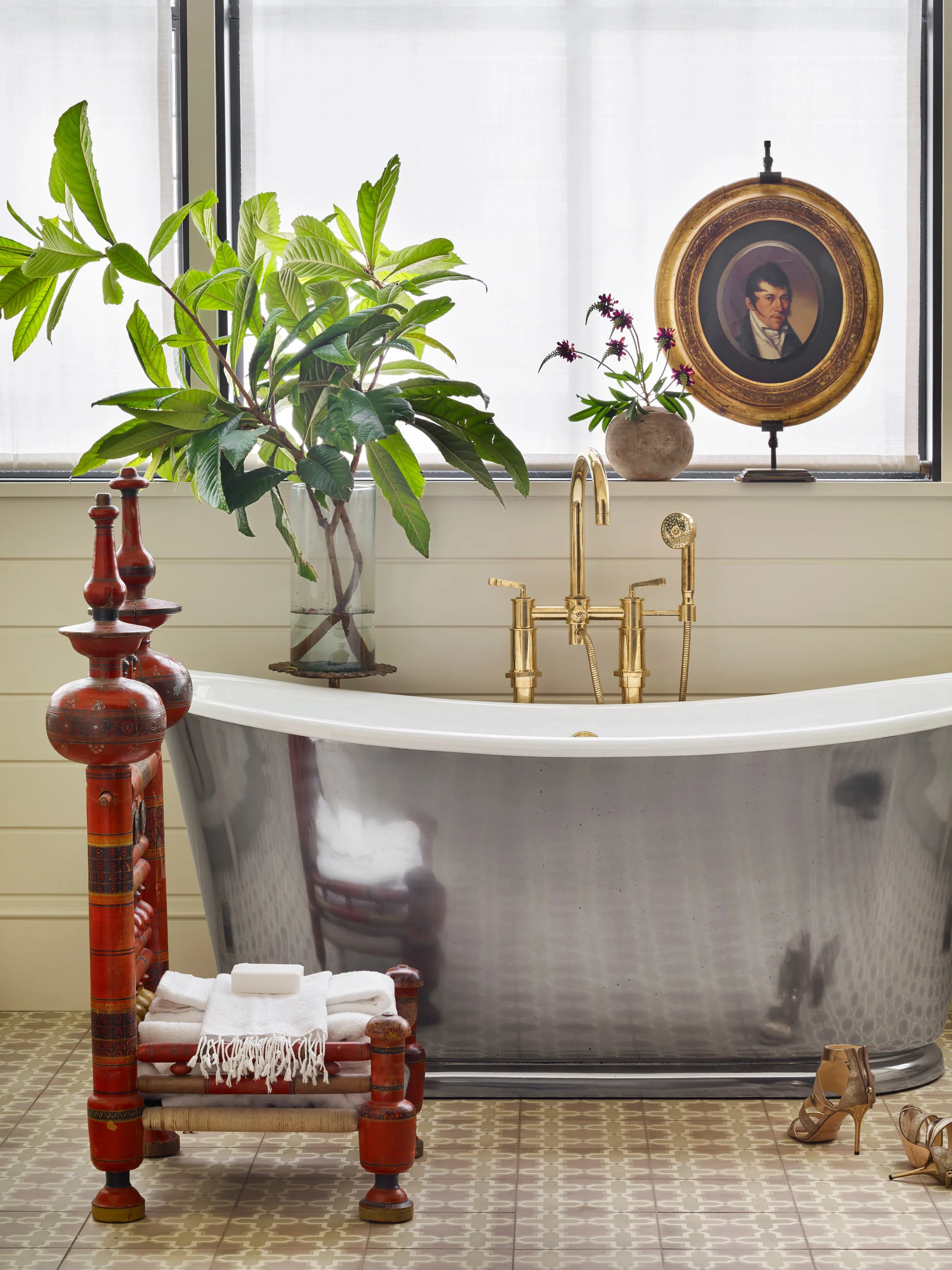 A metallic silver freestanding bathtub sits on patterned tile flooring in front of a window, accompanied by a red decorative stool holding towels.