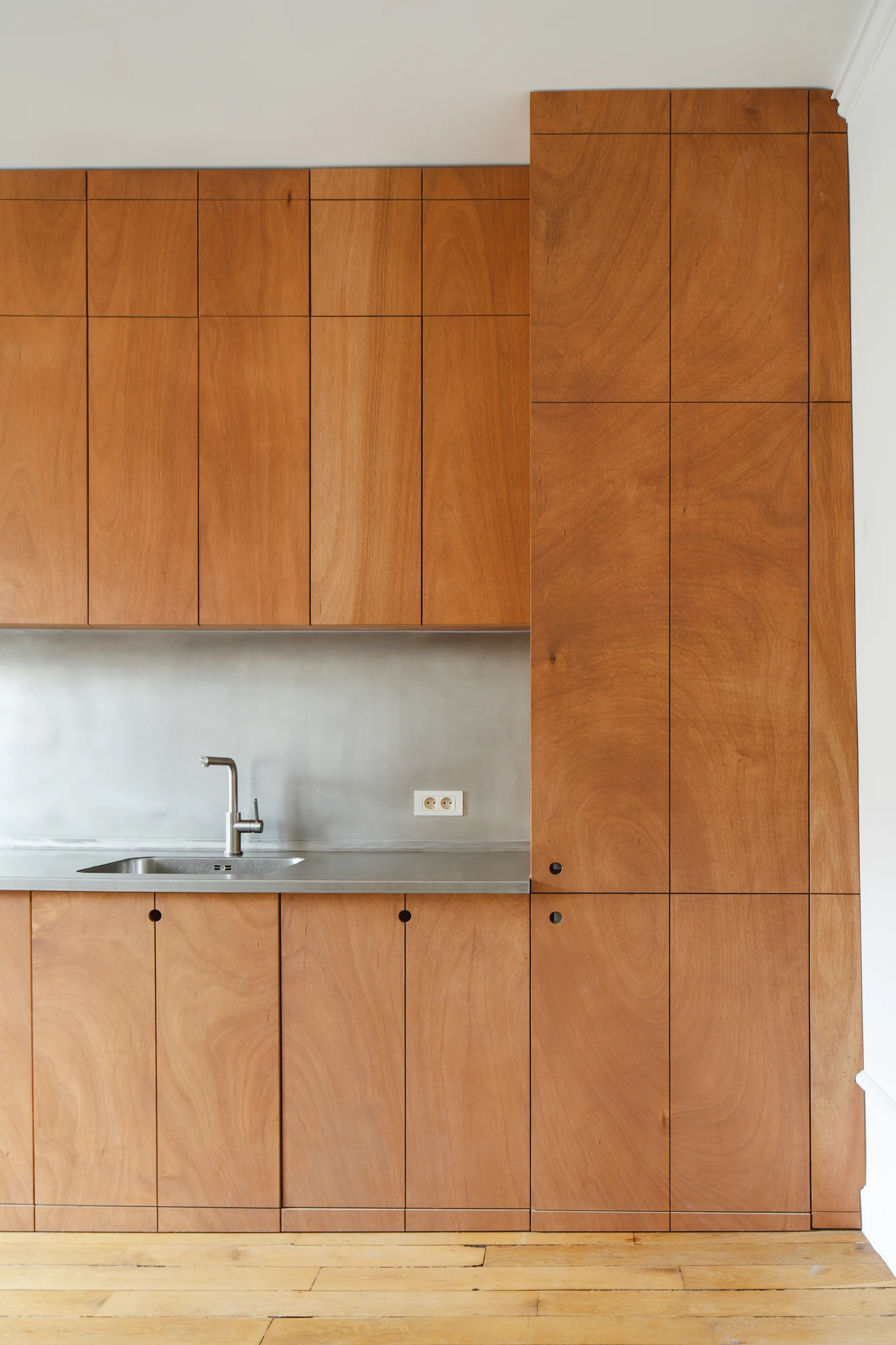 Minimalist kitchen features warm wood grain cabinets with small circular cutout handles alongside a stainless steel countertop, sink, and backsplash.