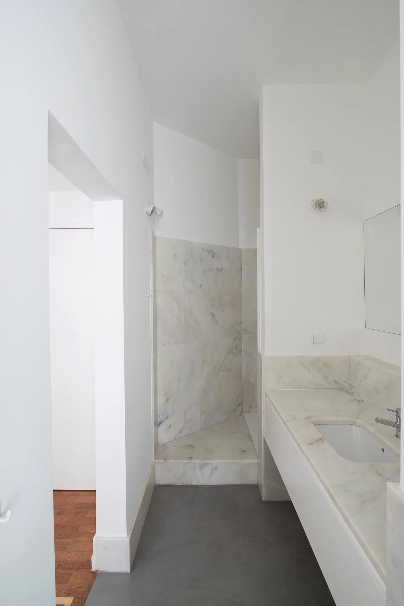 A minimalist bathroom featuring a white marble vanity with integrated sink, a walk-in shower lined with veined marble slabs, and a smooth gray floor.