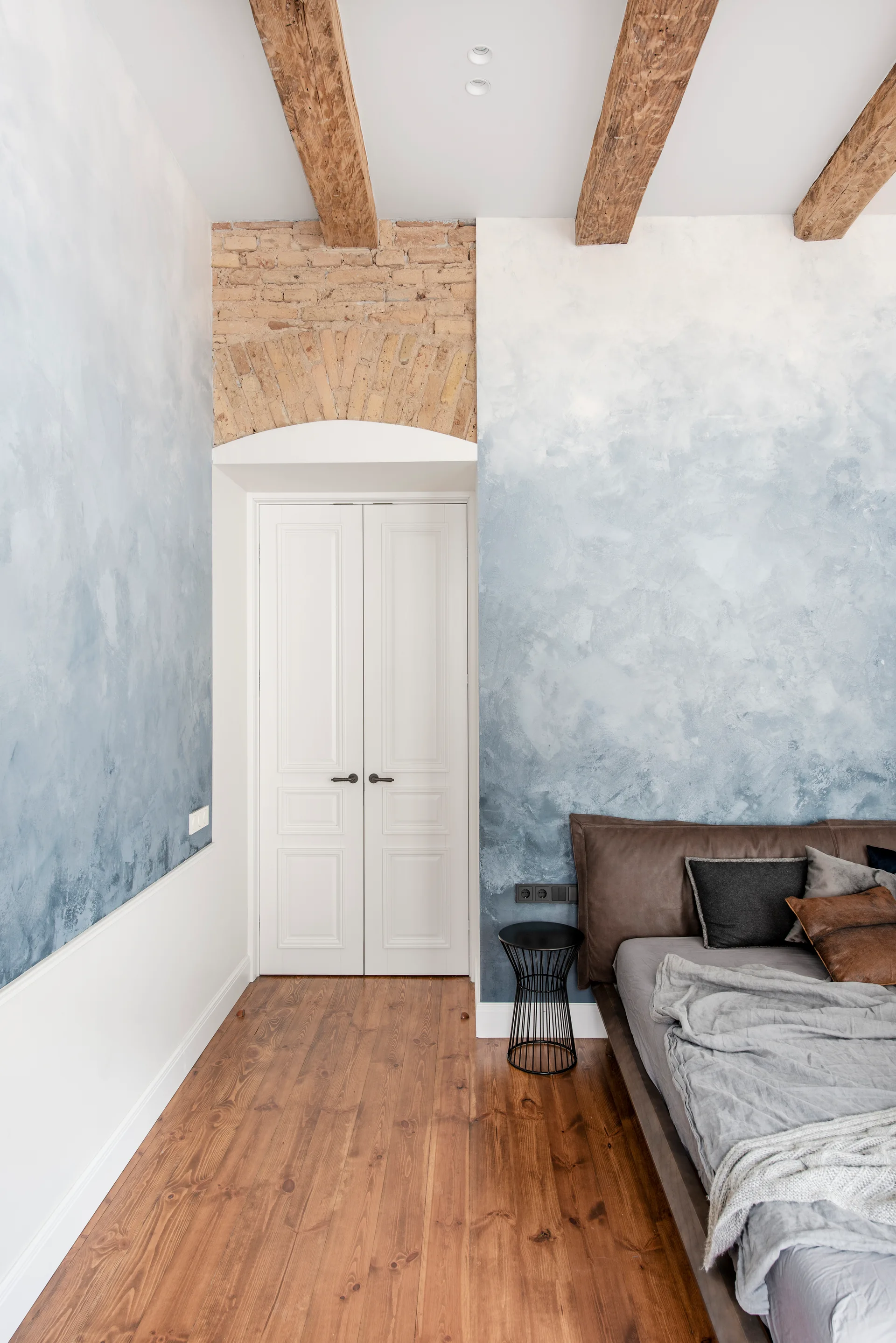A bedroom features textured blue ombre walls, a white double door under a brick arch, and exposed wooden ceiling beams.