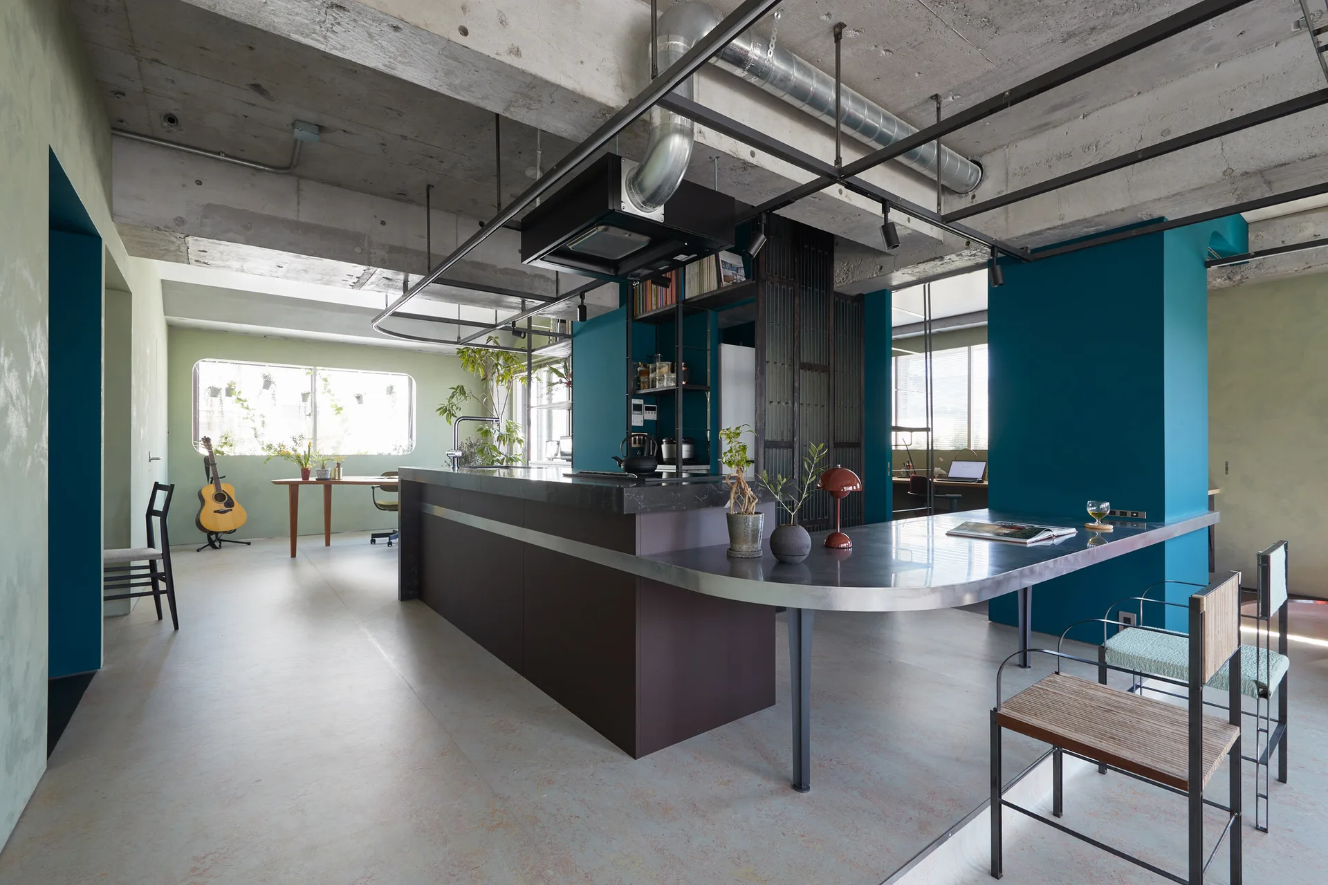 An industrial kitchen features a dark island with an integrated metal dining table under a raw concrete ceiling and vibrant teal accent walls.