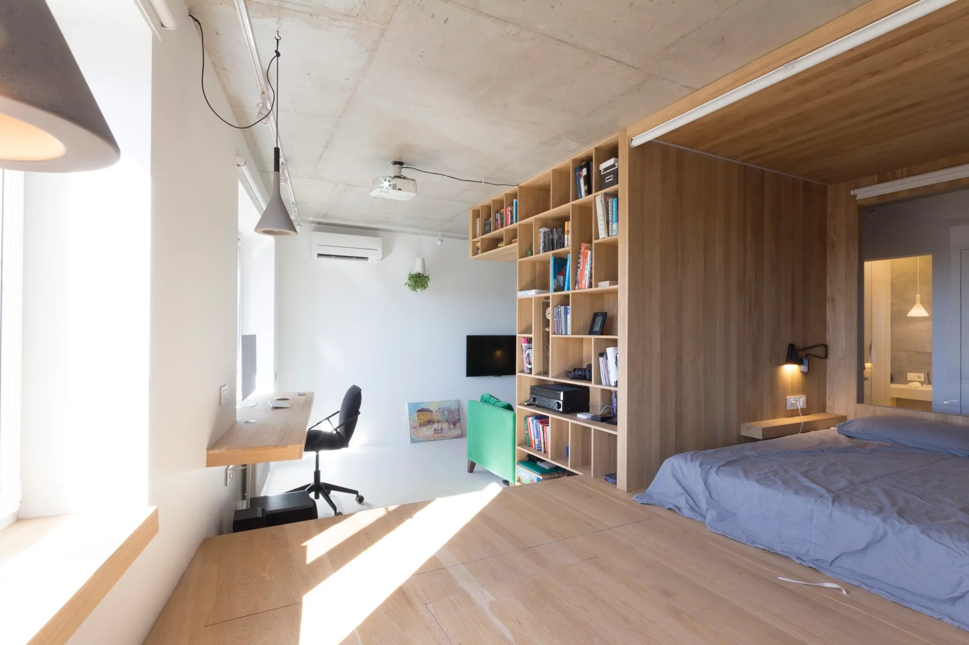 A studio apartment features a raised wooden platform bed, integrated light wood shelving, and a wall-mounted desk beneath a raw concrete ceiling.